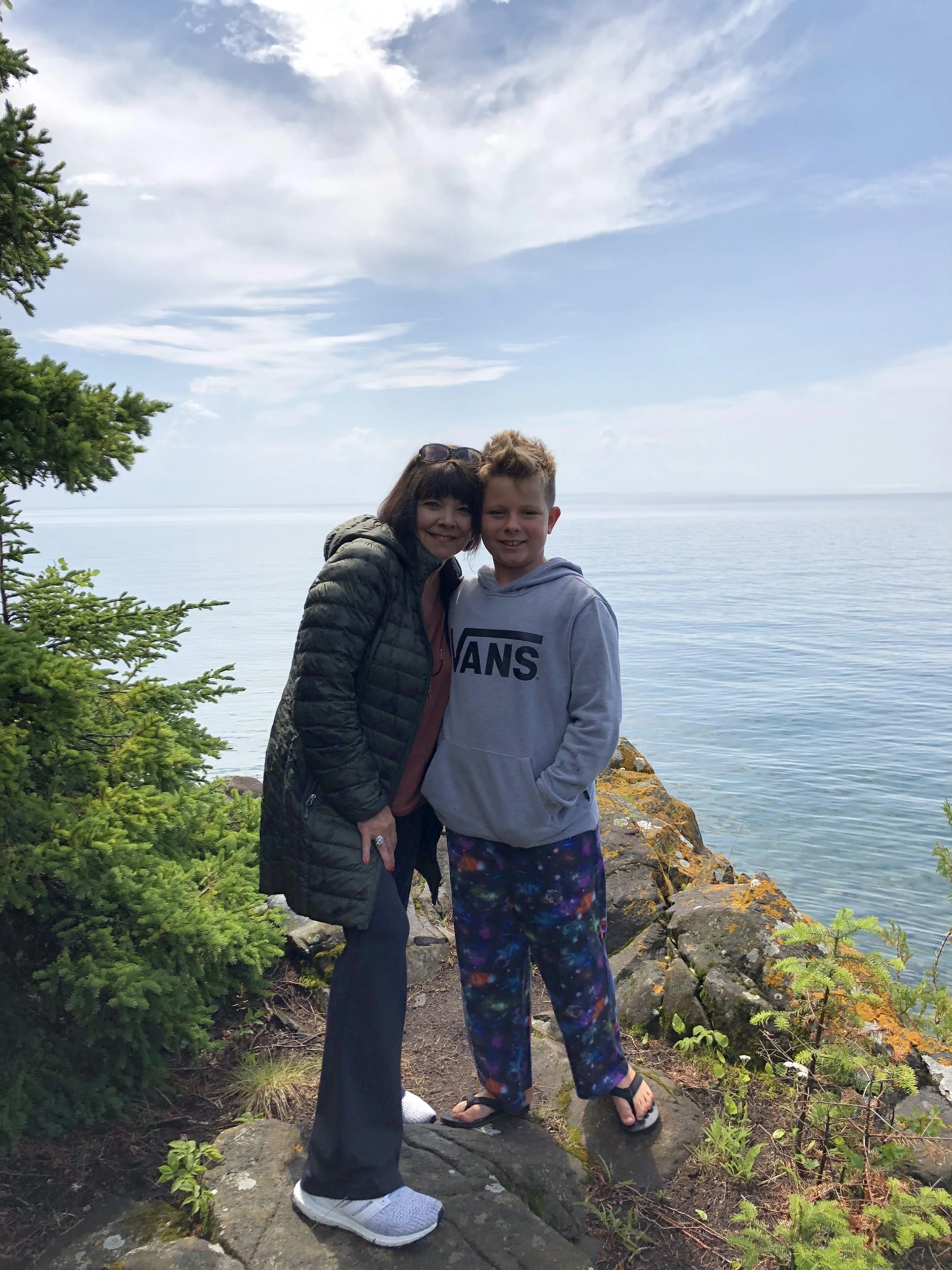 A woman and boy standing on rocks near a body of water with a cloudy sky, greenery, and trees around them.