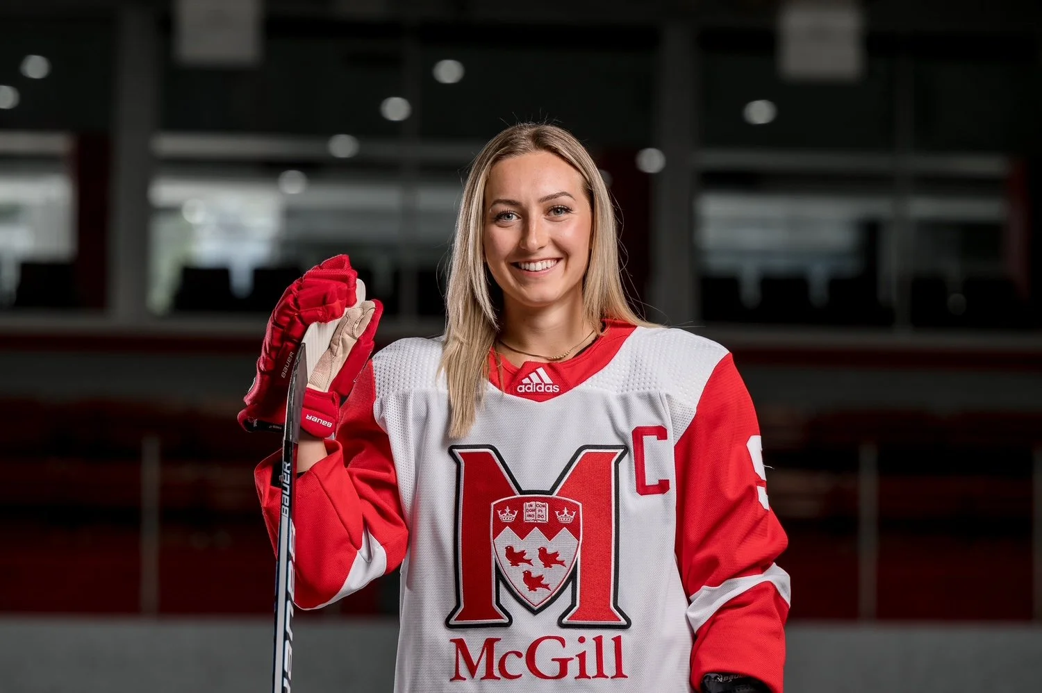 A woman with shoulder-length black hair smiling outdoors in front of green plants.
