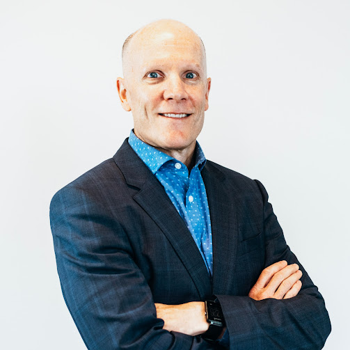 Close-up of a man with curly dark hair, glasses, and a beard, wearing a dark green sweater, looking off to the side with a thoughtful expression.