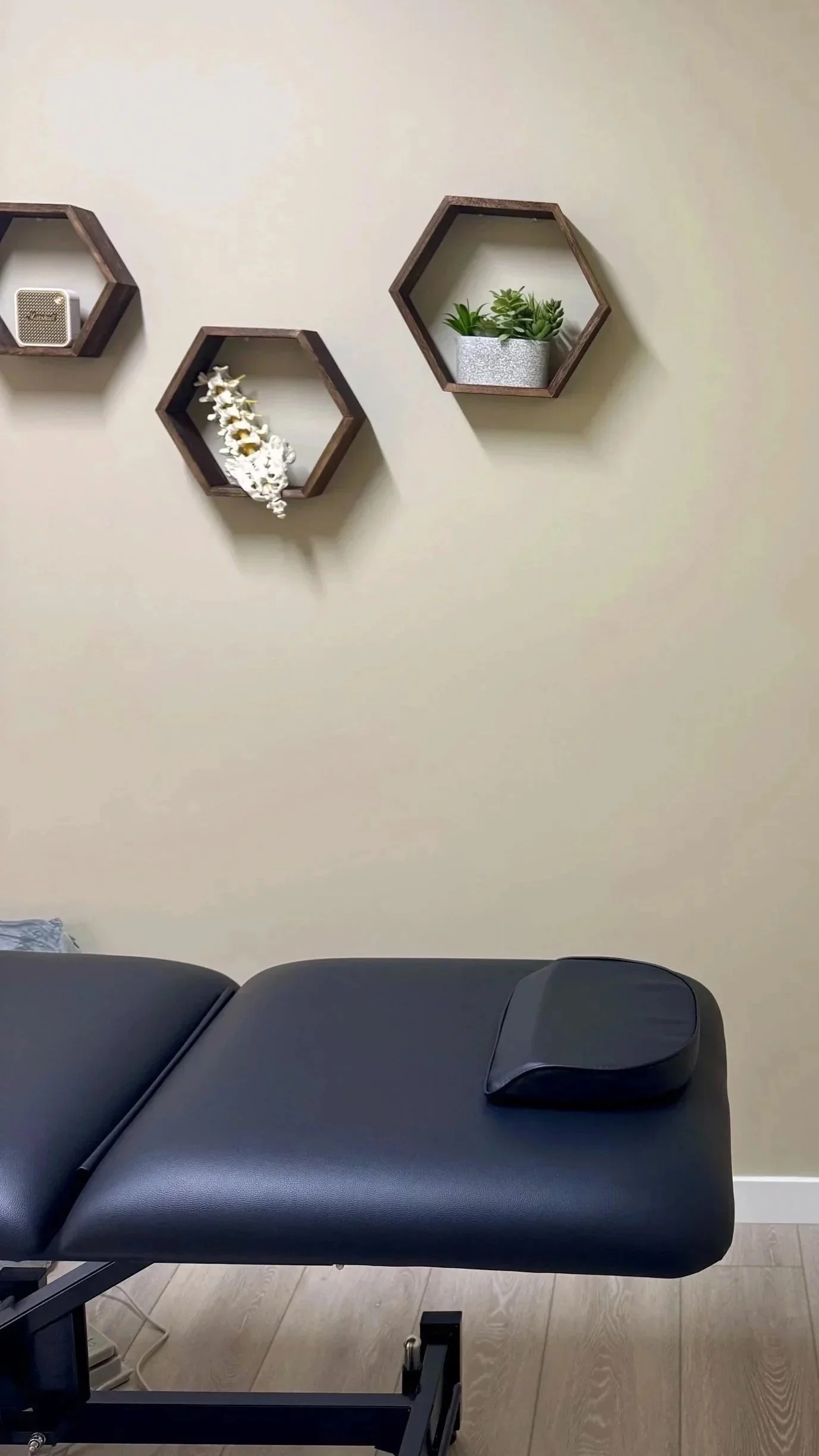 Empty black massage or therapy table in a room with off-white walls, decorated with three wooden hexagonal wall shelves holding a small speaker, a white floral arrangement, and a pot of succulents.