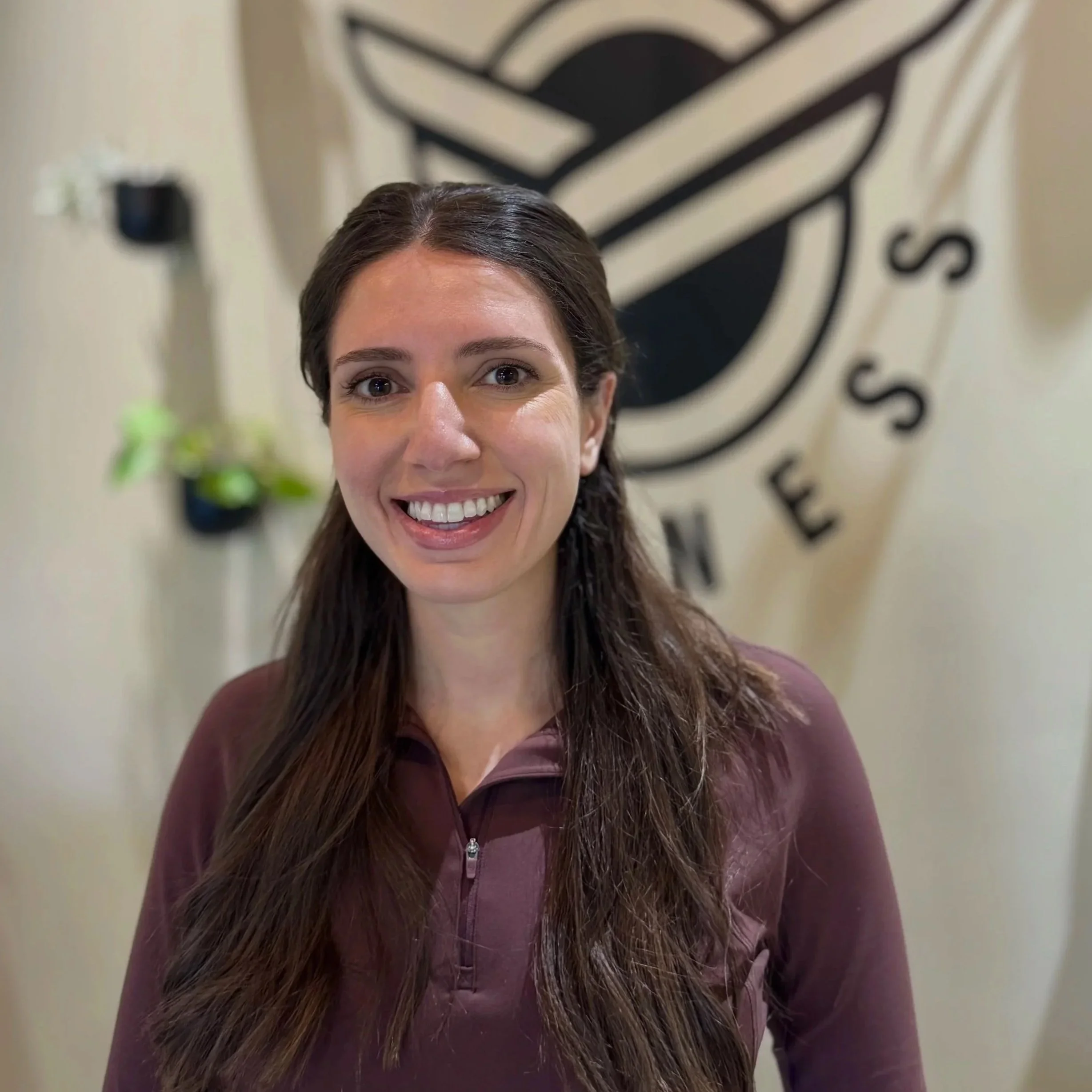 Partial view of a woman with light skin, brown hair, and a friendly expression, standing indoors with a blurred abstract wall art and a plant in the background.