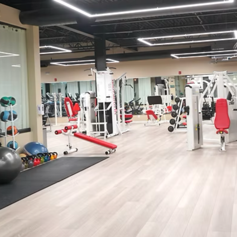 Empty gym with workout equipment including weights, machines, and exercise balls, with hardwood floors and modern lighting.