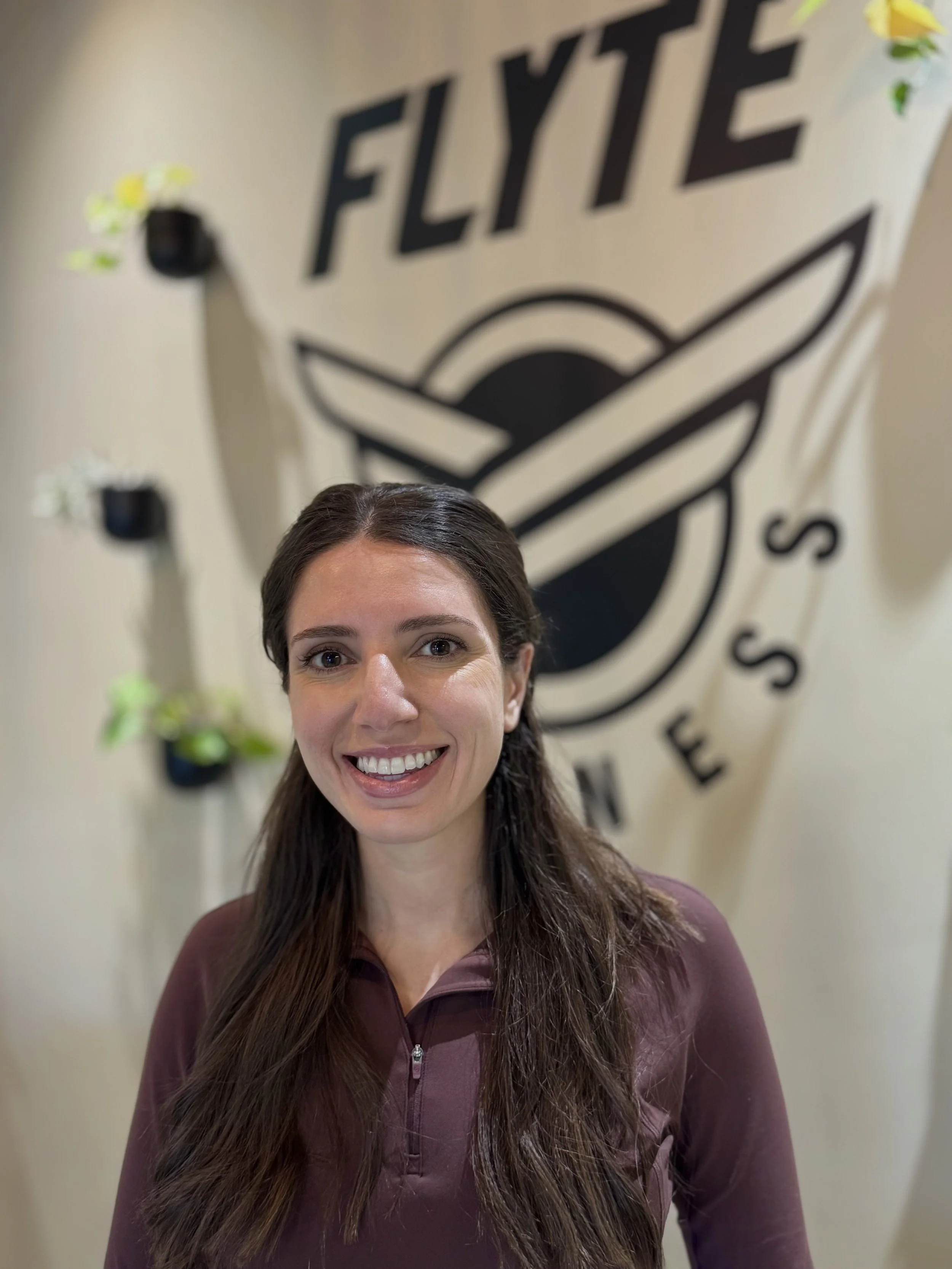 A woman with long brown hair smiling in front of a wall with a logo that says 'FLYTE' and has a stylized airplane design, with some decorative plants nearby.