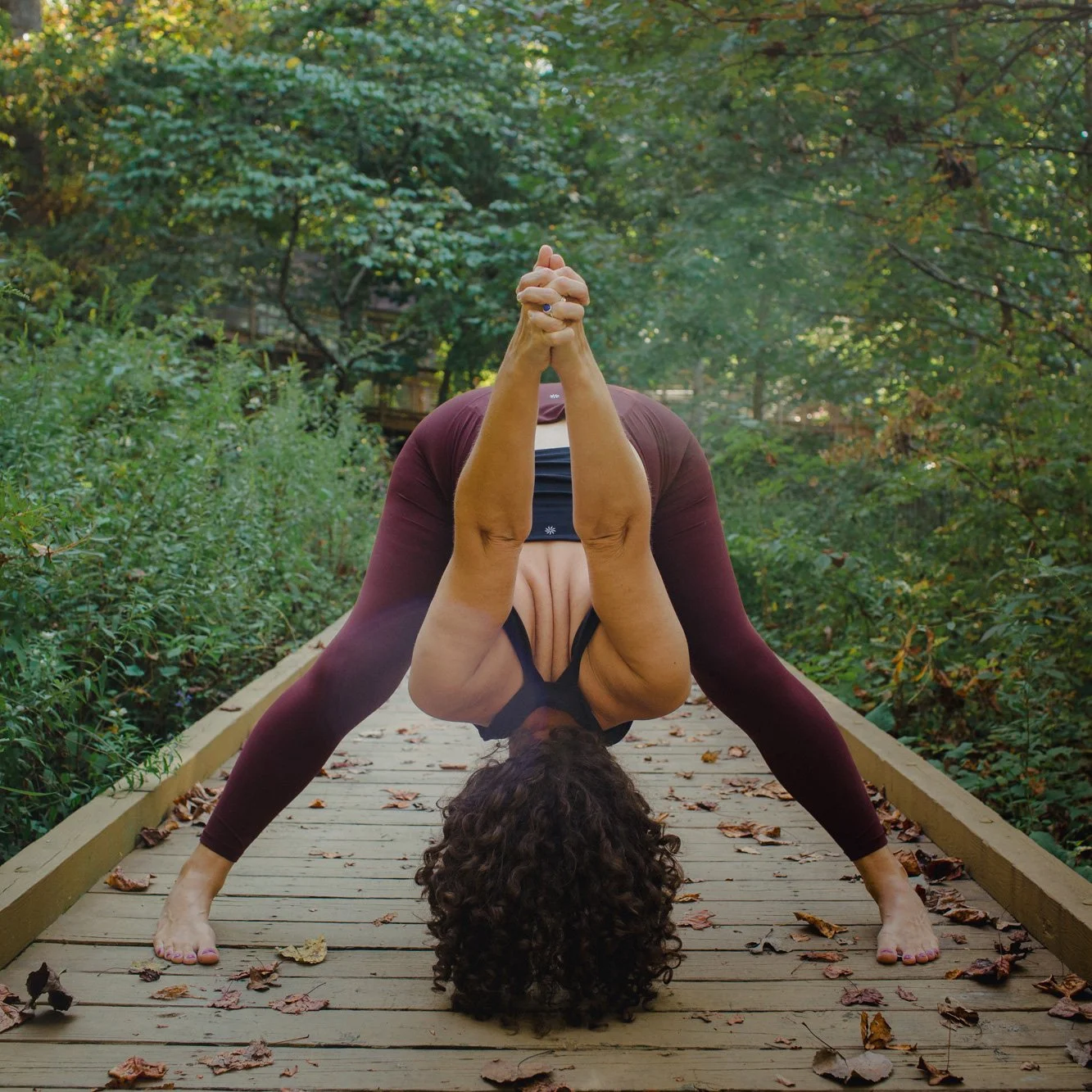 A woman practicing yoga on a wooden bridge surrounded by trees and fallen leaves, bending forward with hands clasped above her head.