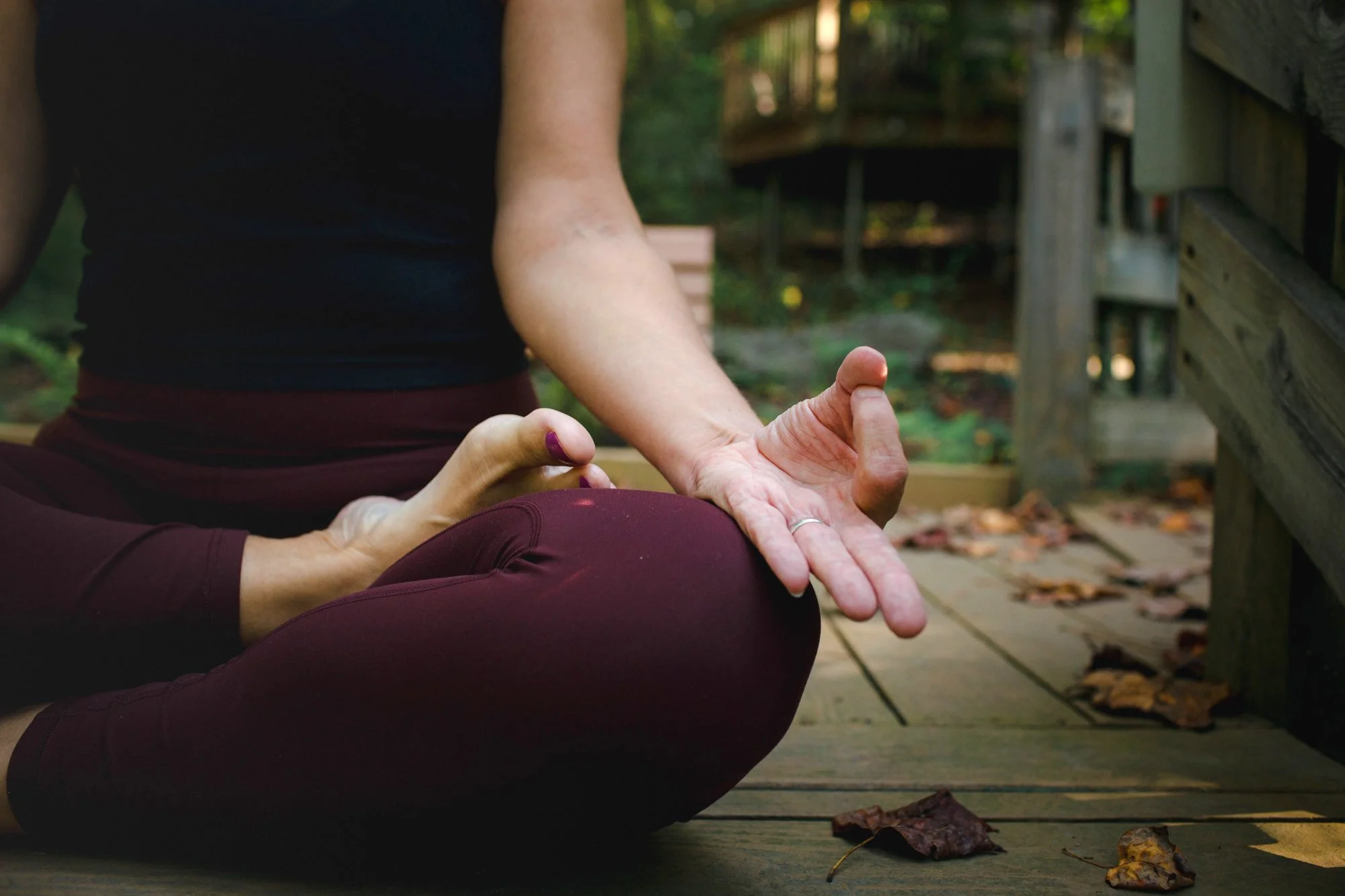 Person practicing yoga outdoors on a wooden deck, sitting cross-legged with one hand resting on their knee and the other forming a mudra with thumb and index finger, surrounded by fallen leaves and lush greenery.