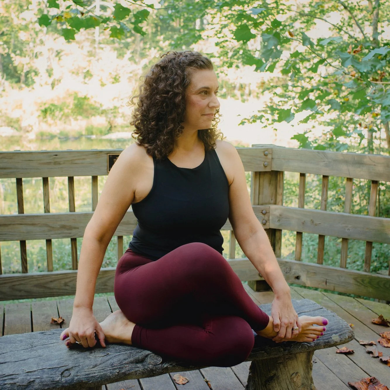 Woman practicing yoga outdoors on a wooden bench, surrounded by green trees and fallen leaves.