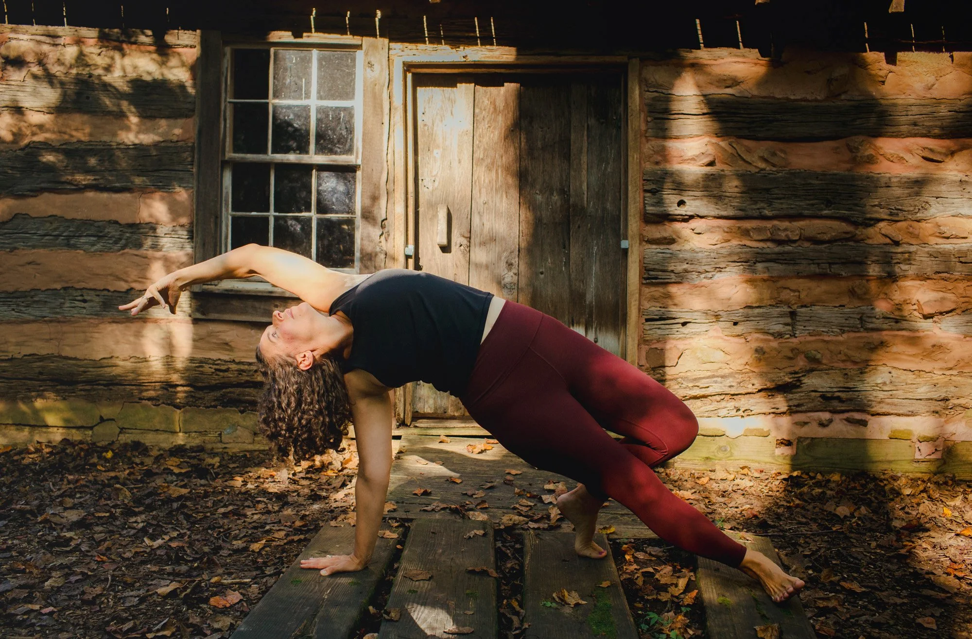 A woman with curly hair practicing yoga outside on a wooden platform surrounded by fallen leaves, in front of an old rustic cabin with a wooden door and window. She is in a side plank pose with one arm reaching overhead.