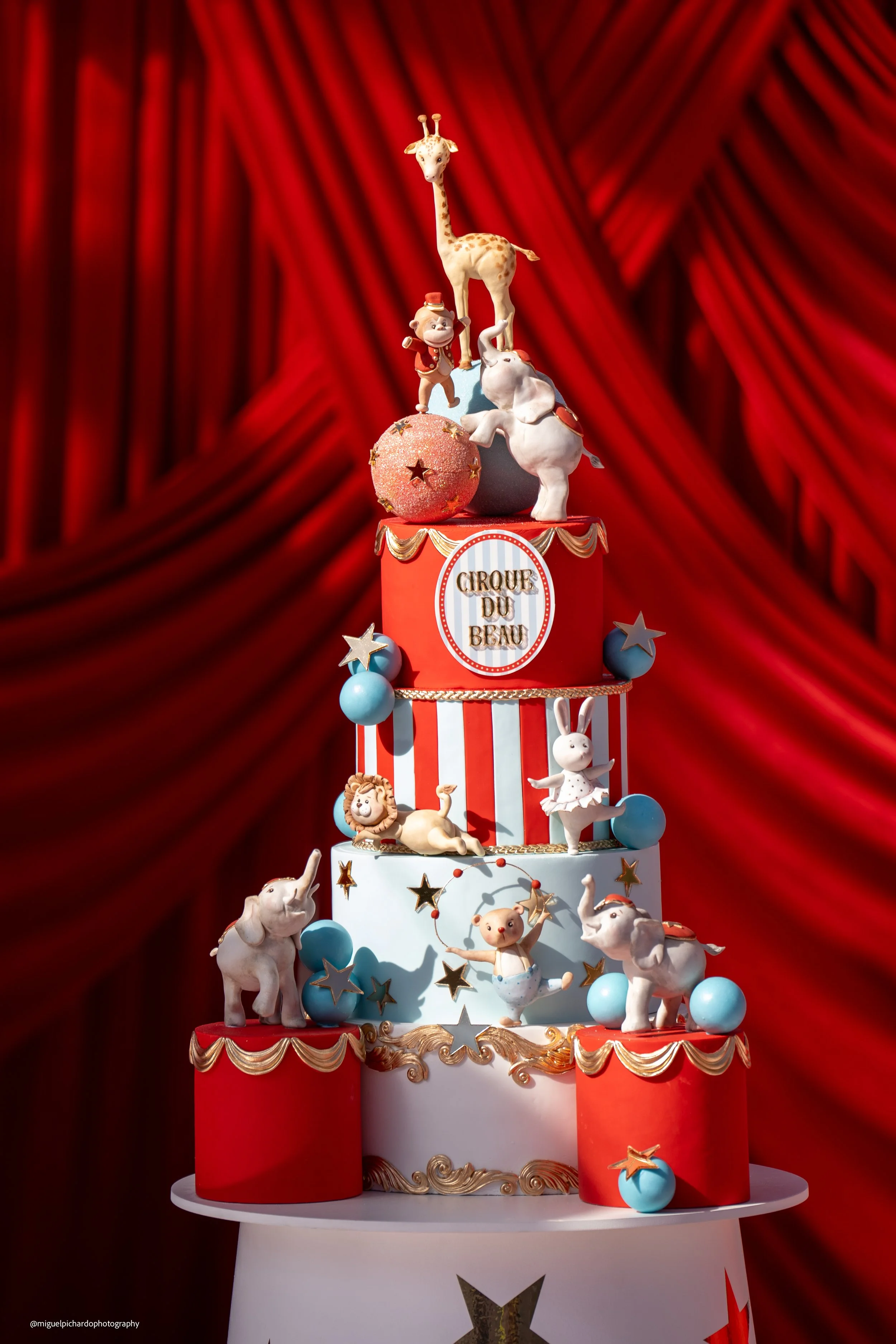A multi-tiered circus-themed cake with circus animals and decorations, set against a red curtain background.