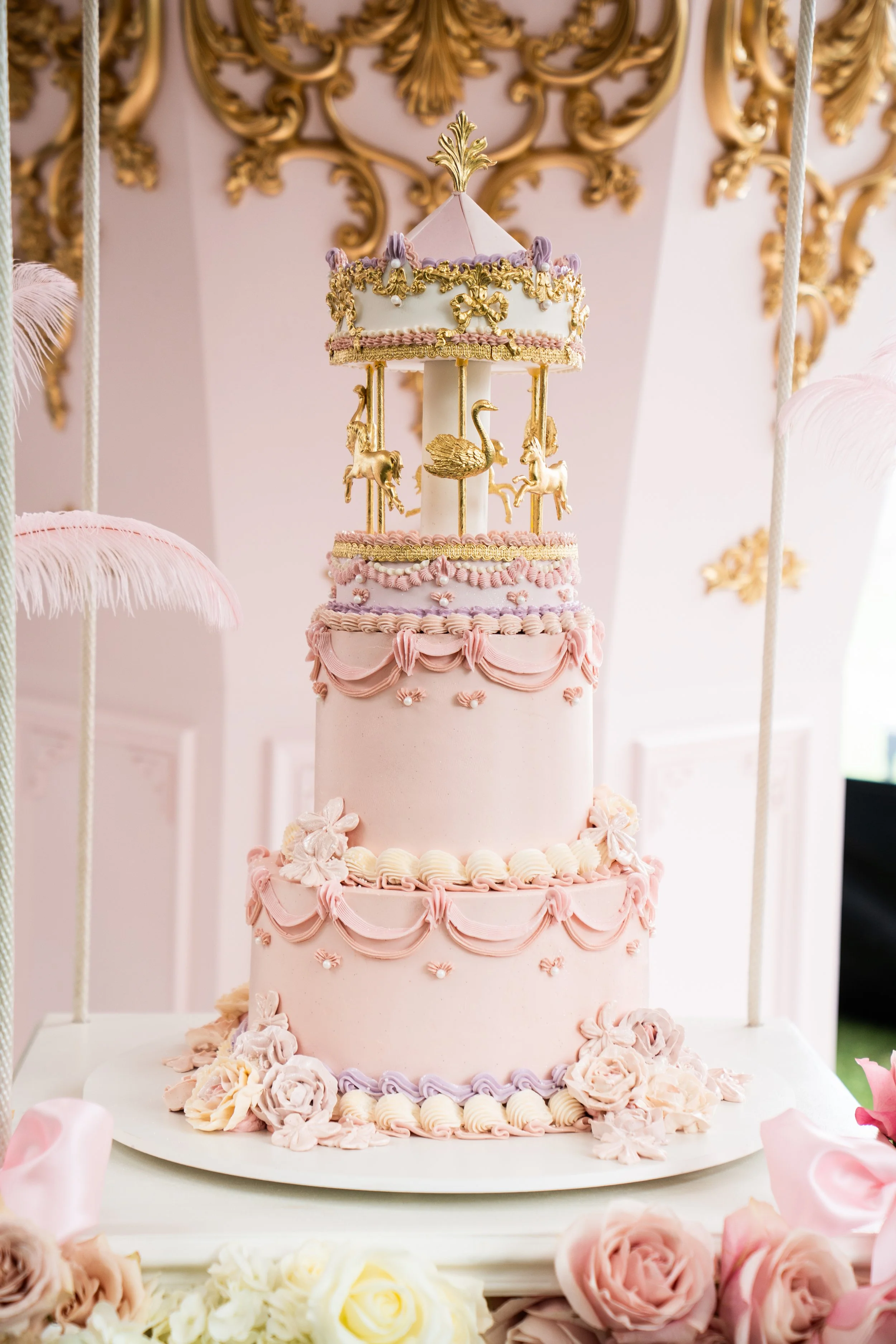 A three-tiered pink cake decorated with pink and lavender icing, floral decorations, and topped with a carousel featuring gold and pastel-colored animals and a pink canopy.