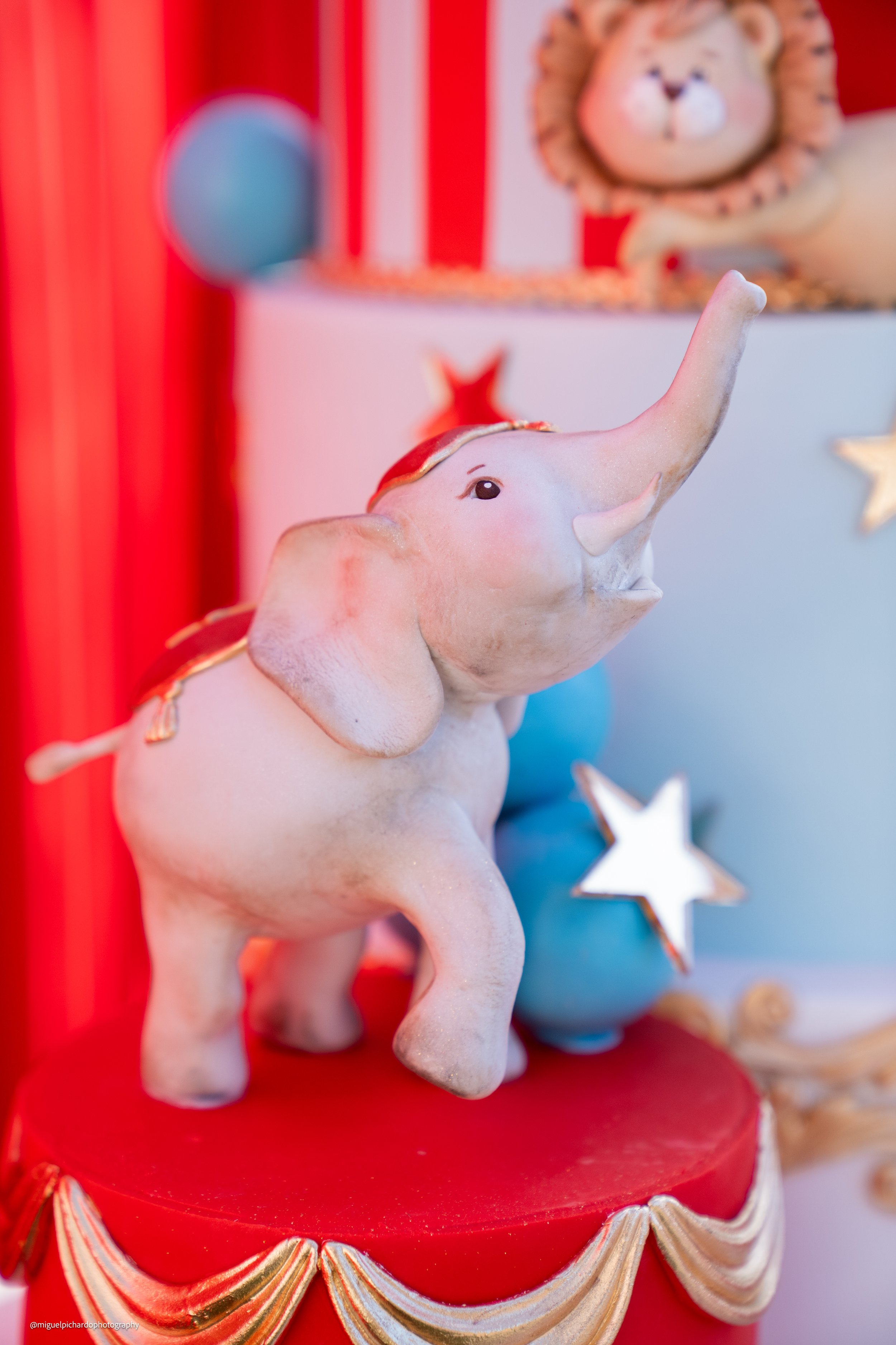 Decorative figurine of a white elephant with a red blanket and gold accents, standing on a red circular surface with gold and white trim. In the background, a blue ball, star, and a lion plush toy are visible, with red and white striped walls.