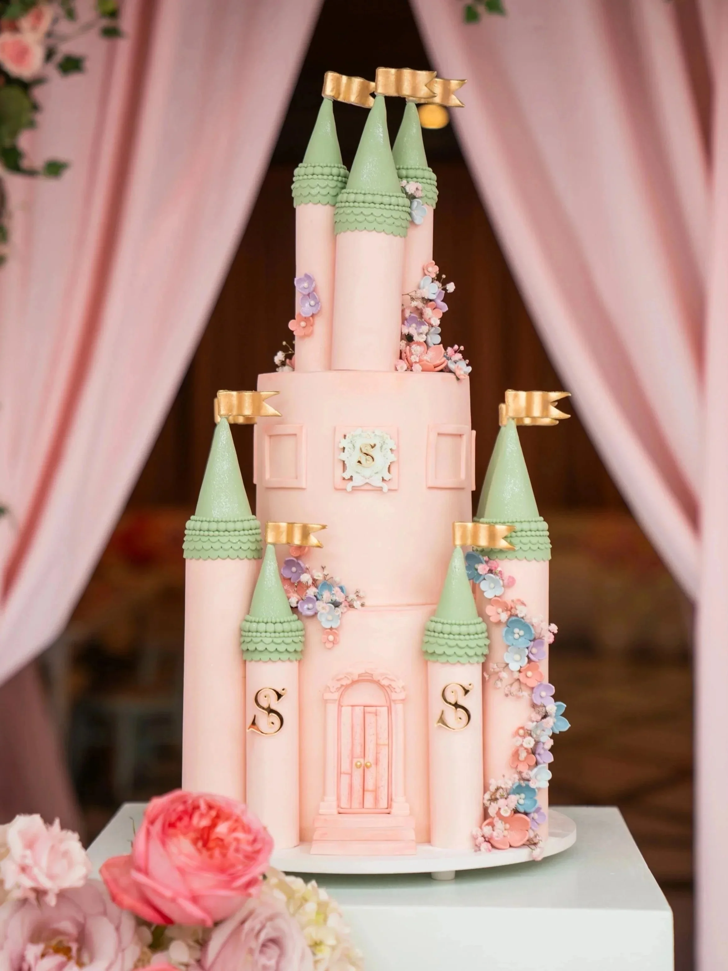 A pink castle-themed cake with green towers and gold flags, decorated with pastel-colored flowers.