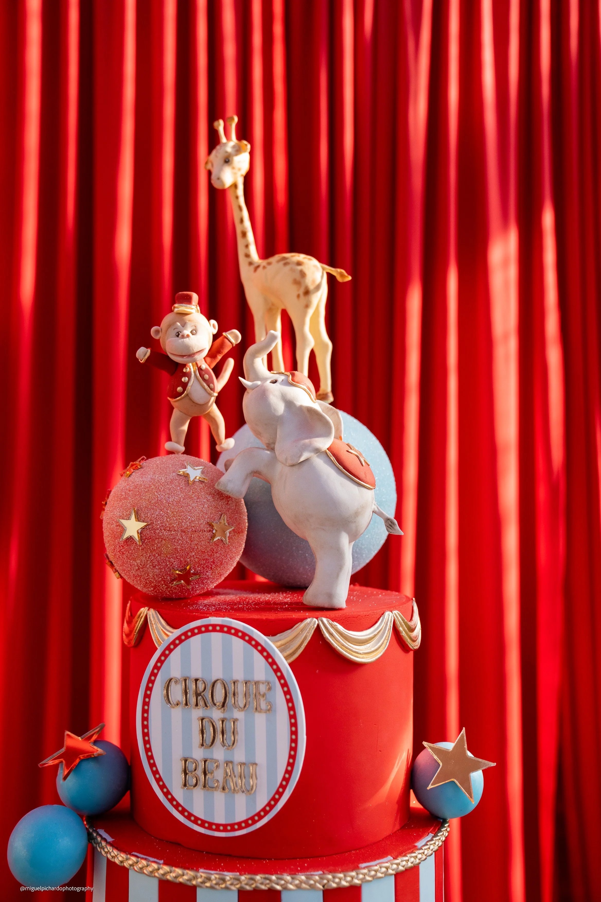A circus-themed cake with circus animal figurines including a giraffe, an elephant, an orangutan, and a monkey, against a red curtain background.