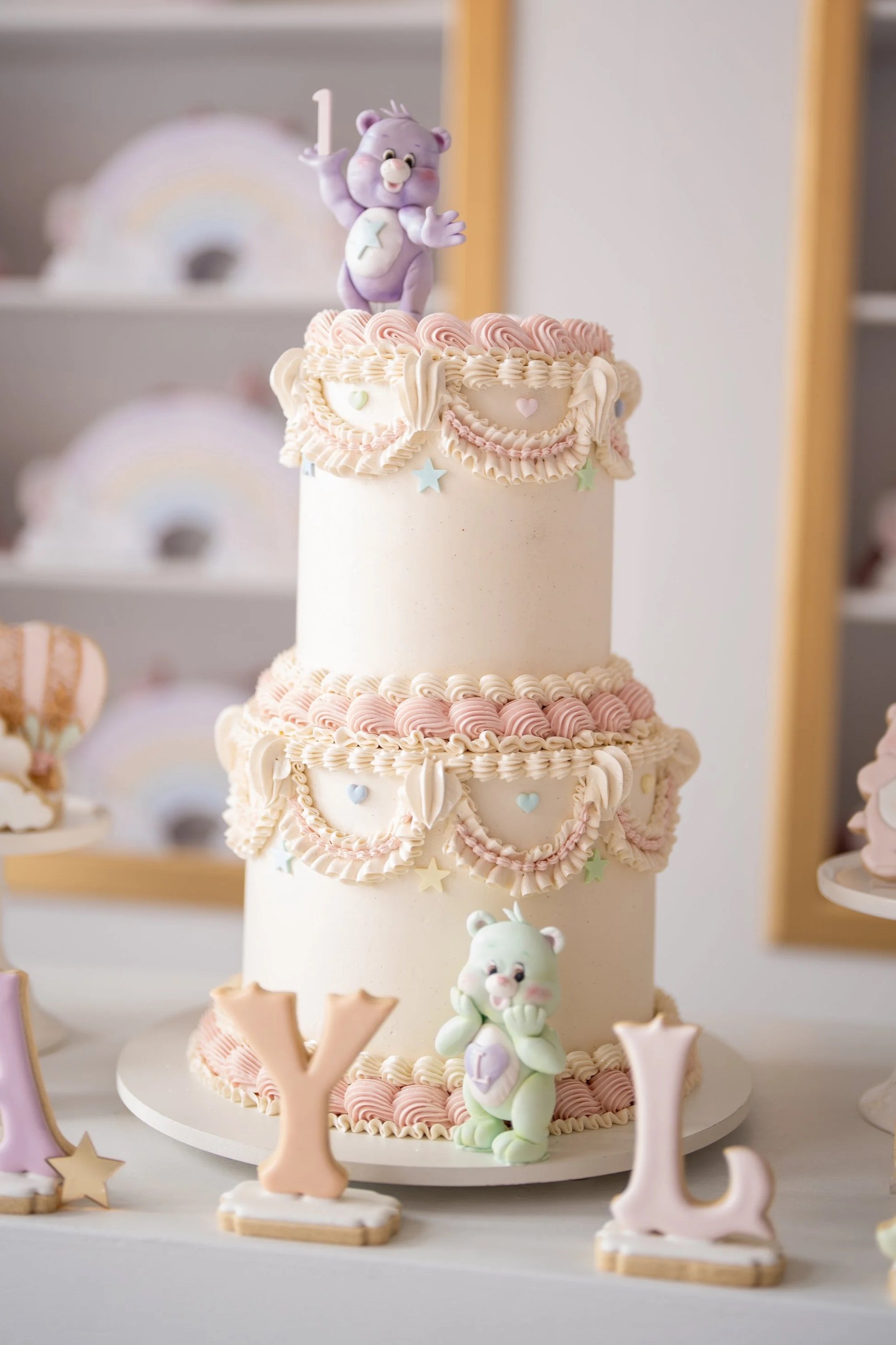 Two-tiered birthday cake decorated with pastel pink and white frosting, star and heart shapes, and edible figurines of a purple bear on top and a white bear at the base. The cake is surrounded by letter cookies spelling 'Y L'.