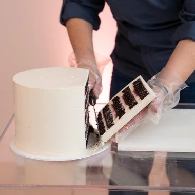 A person in a dark blue shirt is slicing into a layered birthday cake with white frosting, revealing chocolate and vanilla layers inside.