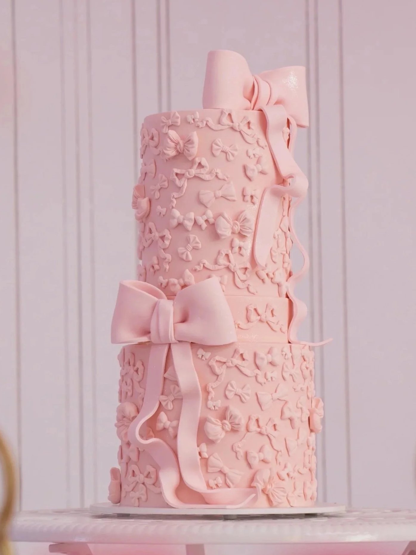 A tall, pink, tiered cake decorated with fondant bows and ribbons, featuring intricate bow-shaped decorations scattered across the surface.