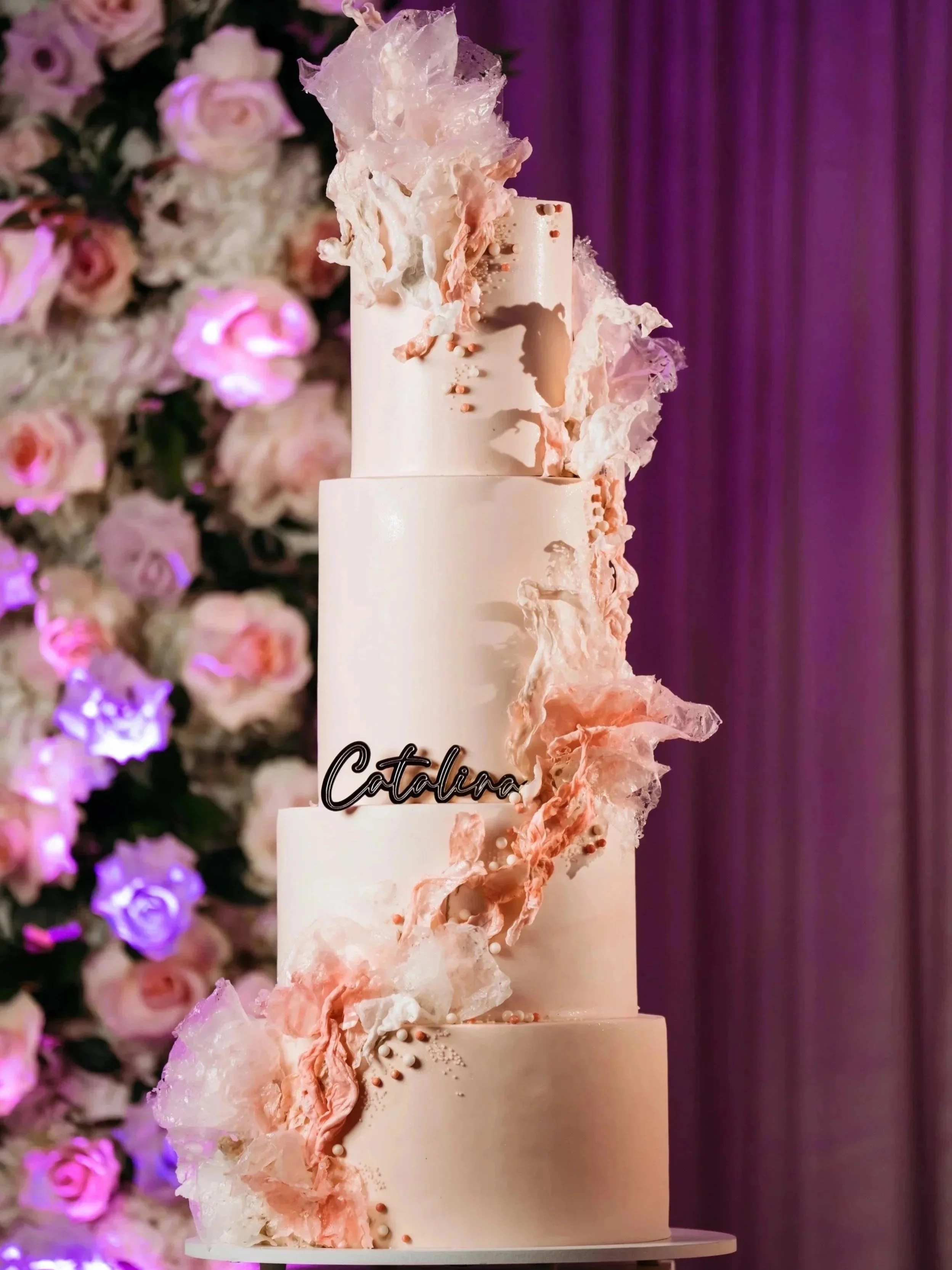 Elegant multi-tiered wedding cake decorated with pink and white edible sugar decorations and a black 'Catalina' cake topper, set against a backdrop of pink and purple flowers and curtains.