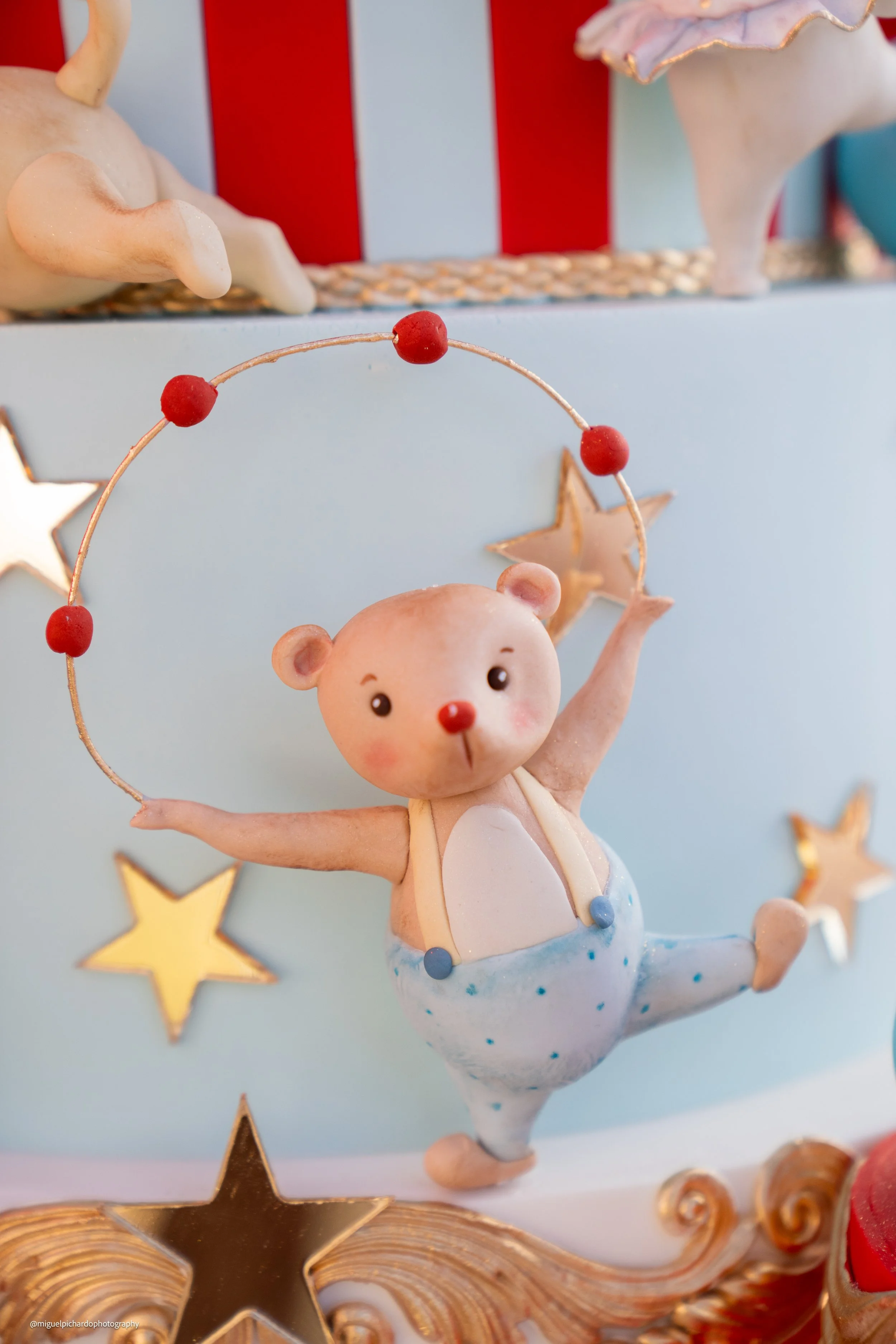A decorative figurine of a dancing bear with a red nose, wearing blue polka dot pants, holding a red-bulb adorned ring, surrounded by star-shaped decorations