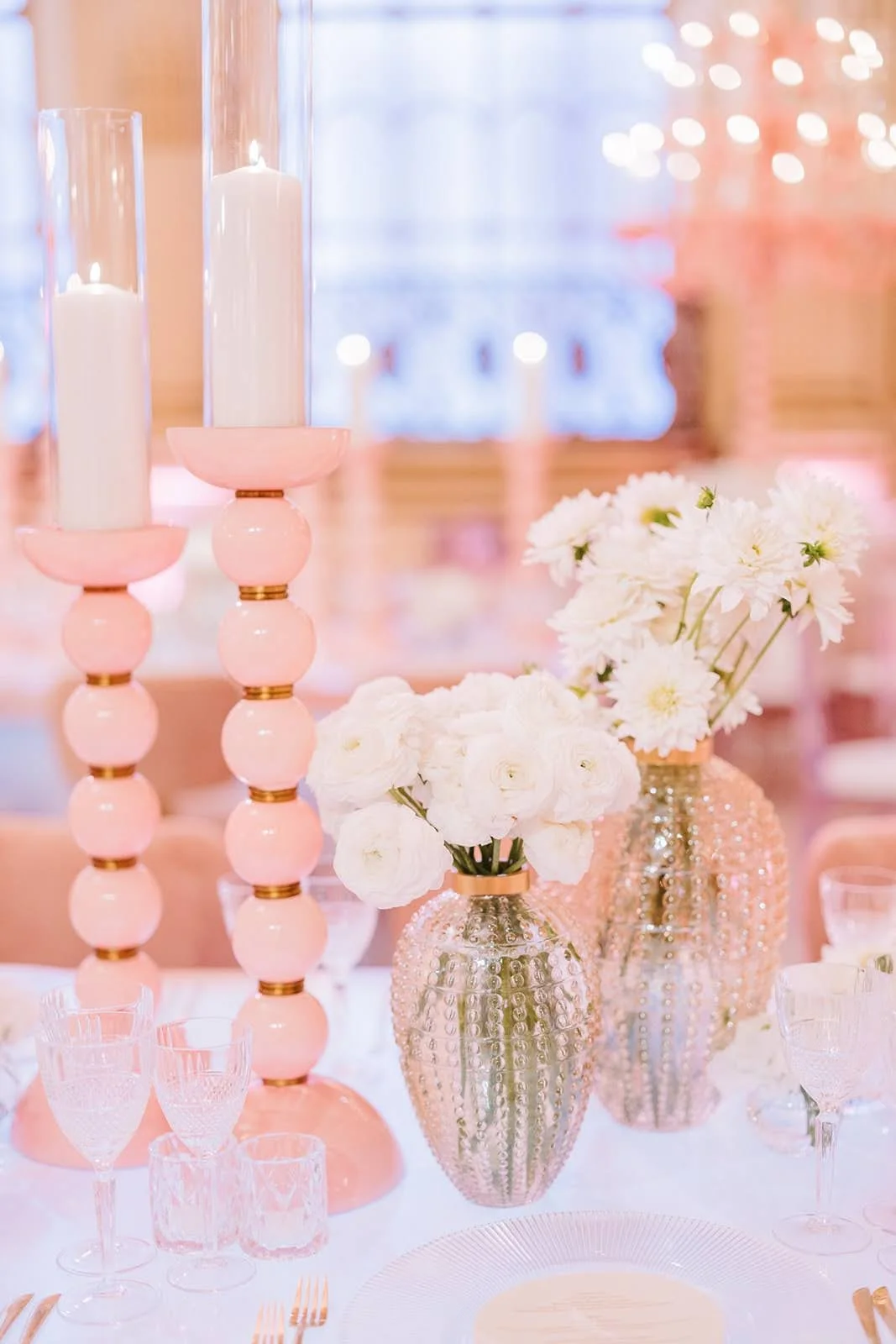 Elegant table setting with pink and white floral arrangements in textured glass vases, tall pink candlesticks with white candles, and glassware, at a softly lit event.