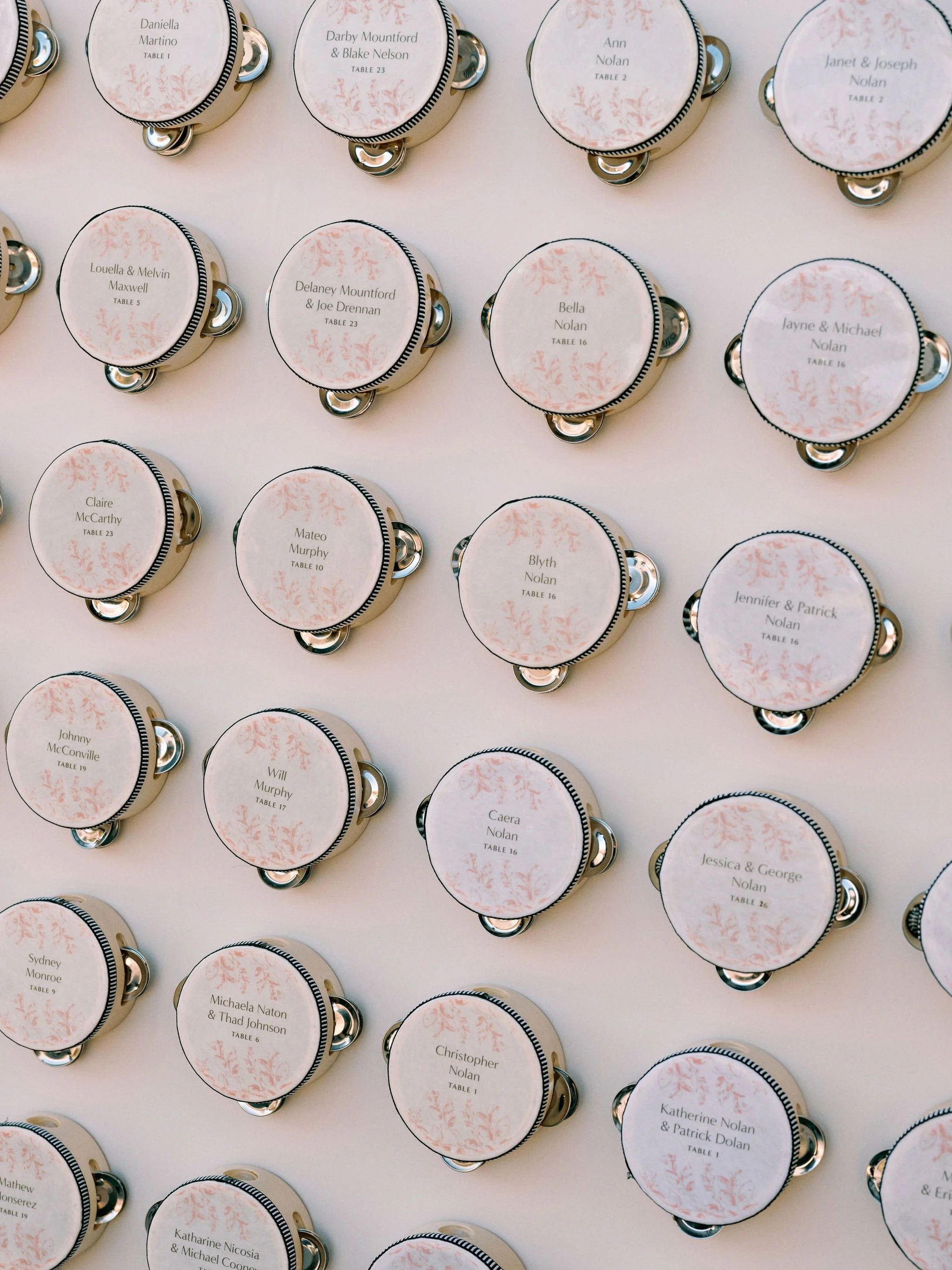 A collection of round table number cards with guests' names for a wedding, arranged on a white surface. Each card displays a guest's name, a table number, and a decorative pink floral pattern.
