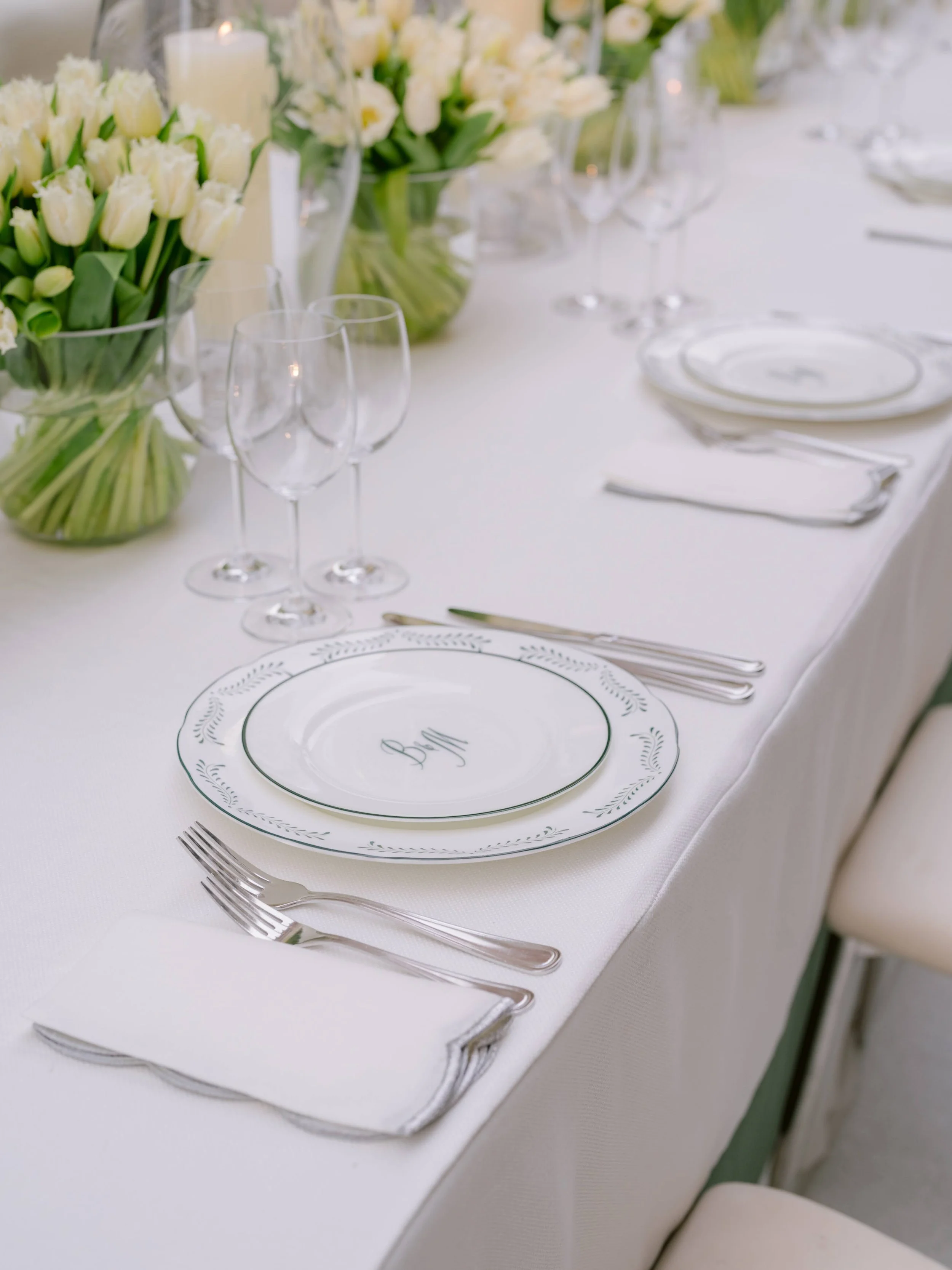 Elegant table setting with white plates, silverware, and wine glasses, decorated with white tulips in vases and candles, for a formal event.