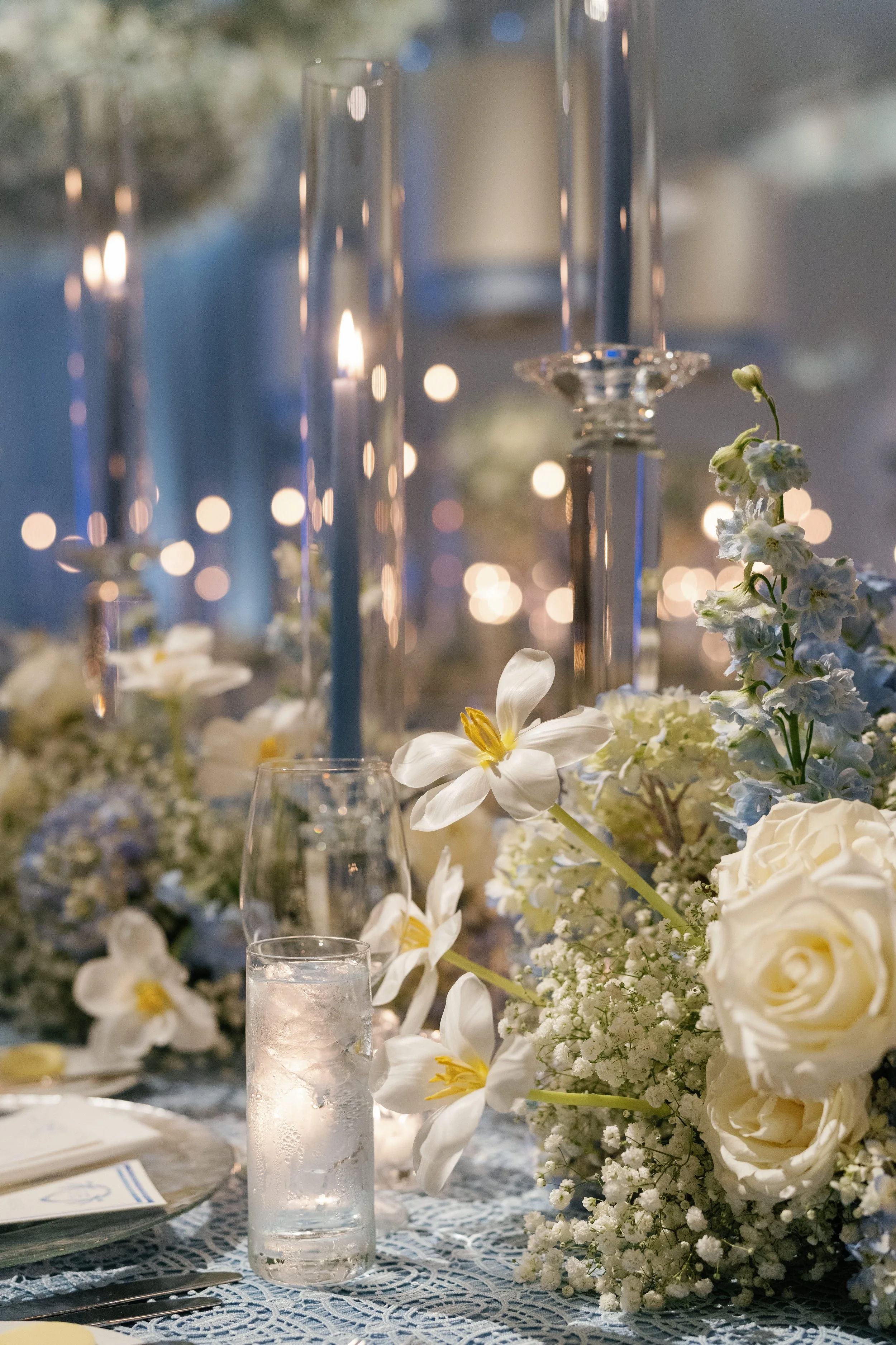 A close-up of a wedding table centerpiece with white flowers, including roses, lilies, and baby's breath, with glass candle holders and tall, thin glass vases, on a lace tablecloth.