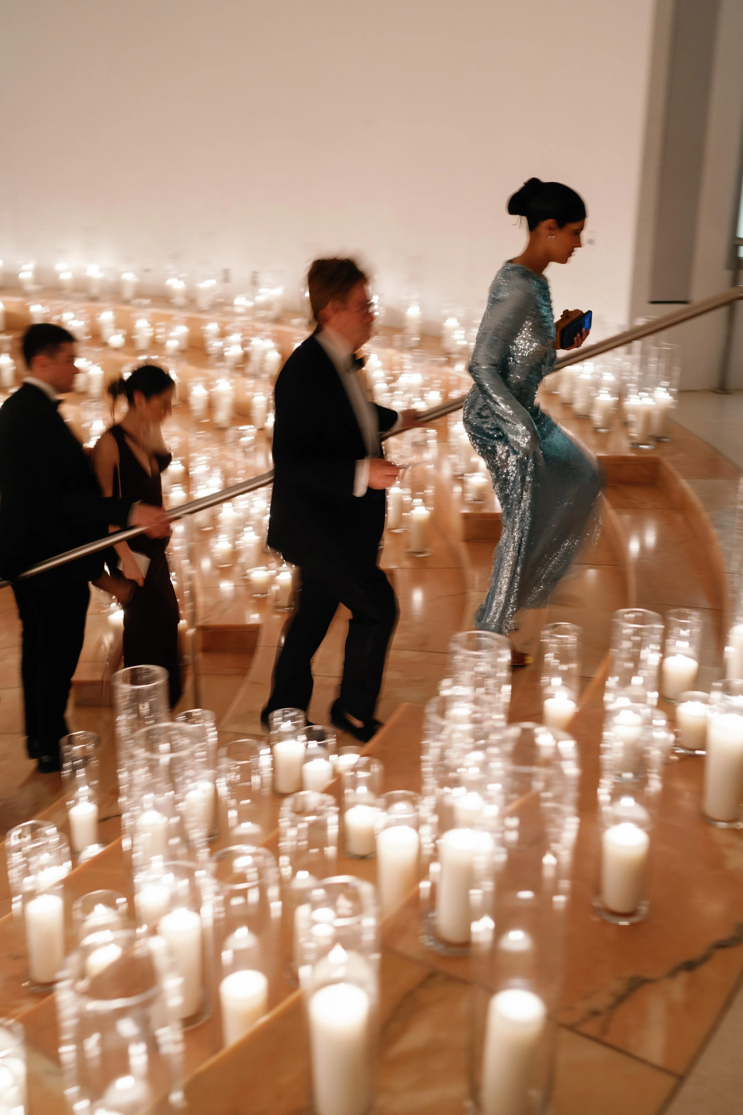 A woman in a sparkling dress is walking up a staircase surrounded by numerous lit candles in glass holders. Four other people follow her, holding the handrail, at a formal event or celebration.
