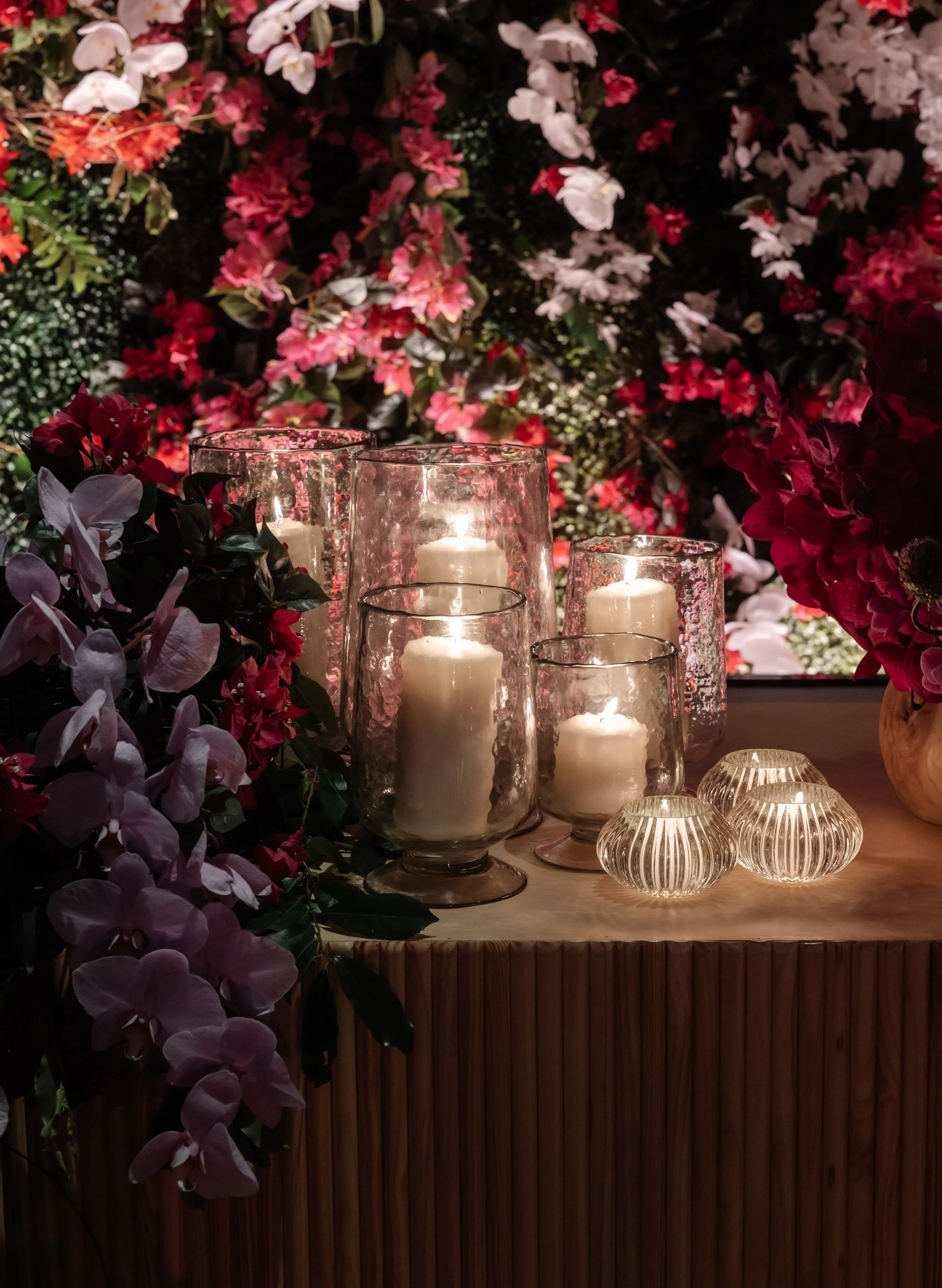 Decorative display of four glass candle holders with white candles on a wooden surface, surrounded by purple and red flowers, with a background of pink and white flowers.