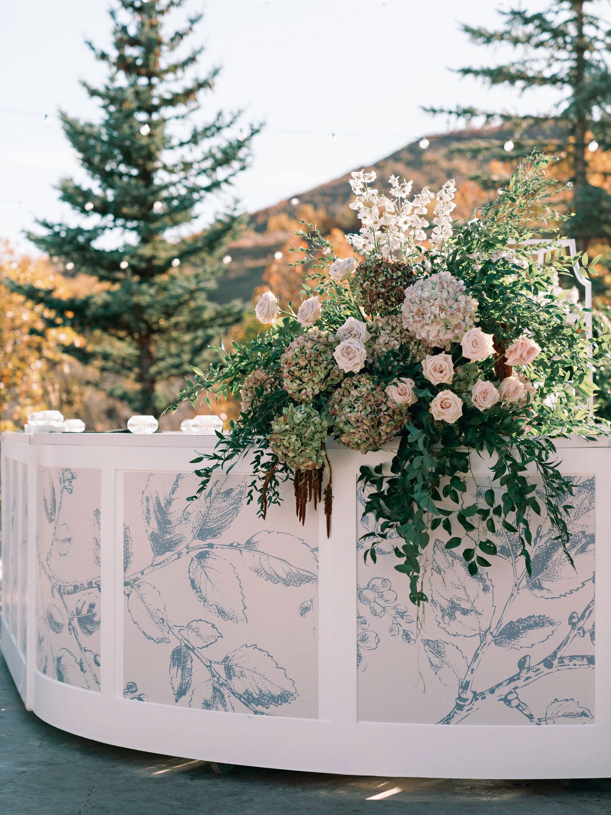 A floral arrangement with pink roses, hydrangeas, and greenery on a white stand with leaf patterns, set outdoors with trees and mountains in the background.