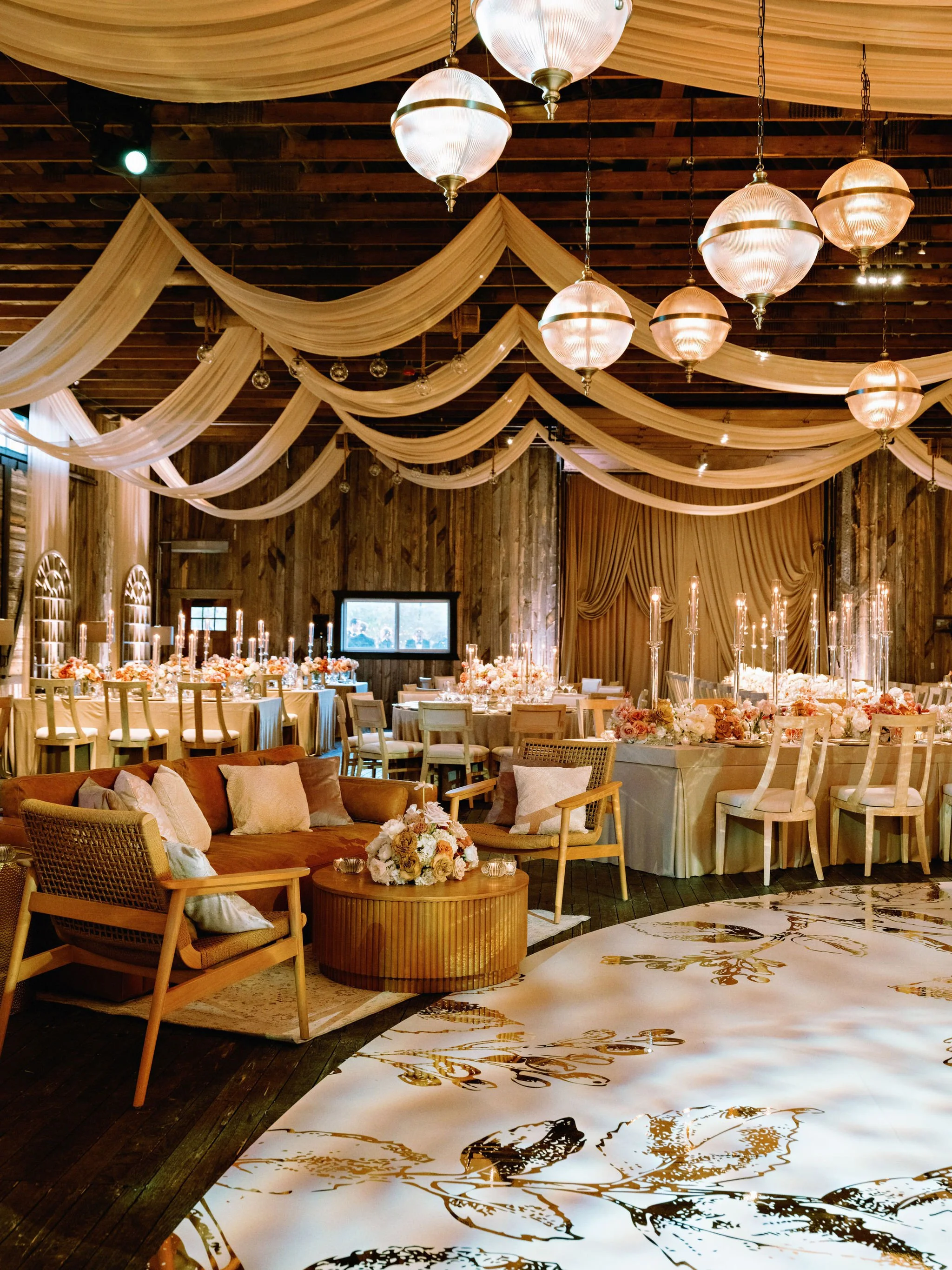 A decorated event hall with draped beige fabric on the ceiling, hanging pendant lights, a wedding-style setup with floral centerpieces, candles, and elegant furniture.