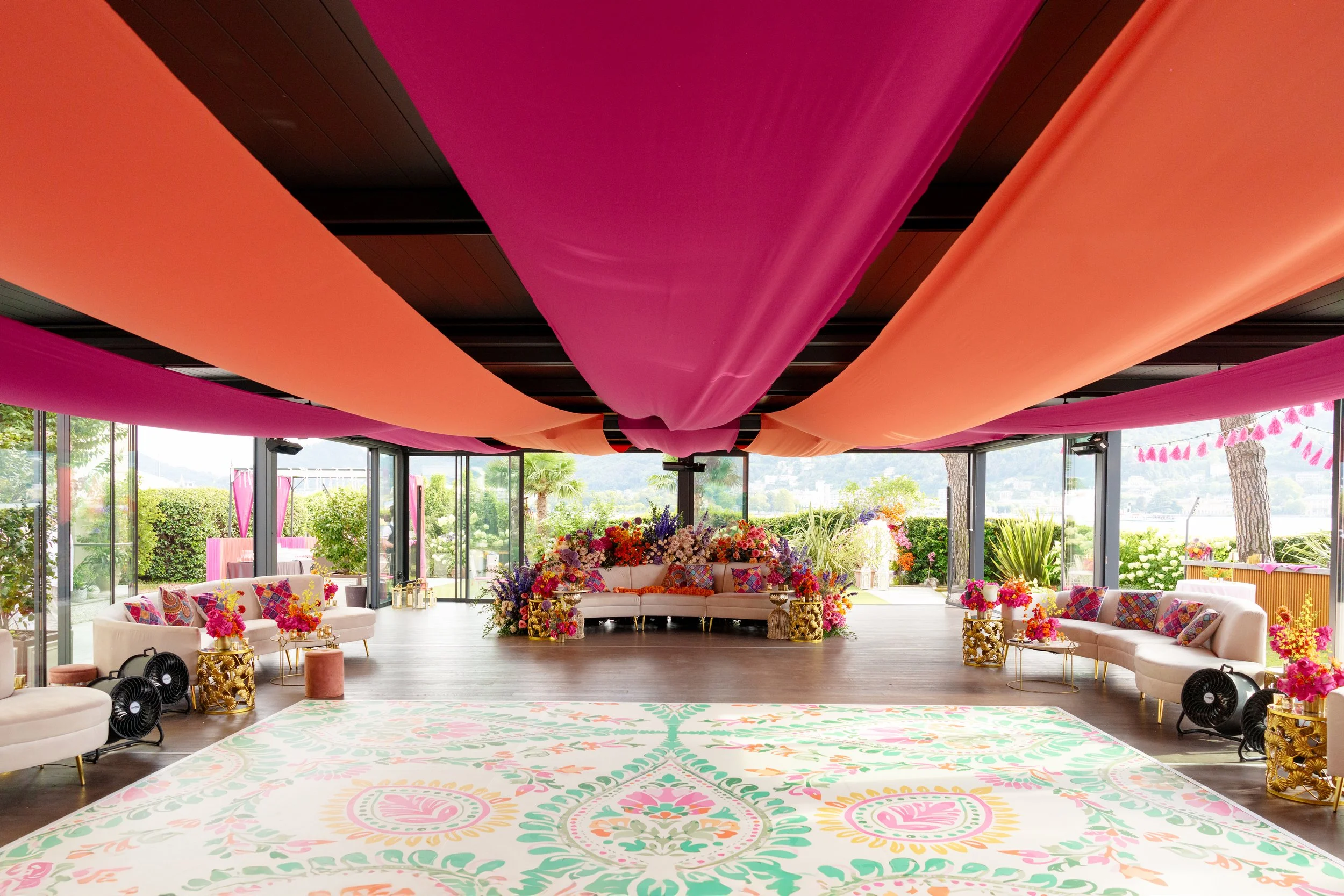 A decorated event space with pink, peach, and orange fabric draped on the ceiling, floral arrangements, white sofas with colorful cushions, and large floor-to-ceiling windows showing an outdoor garden scene.