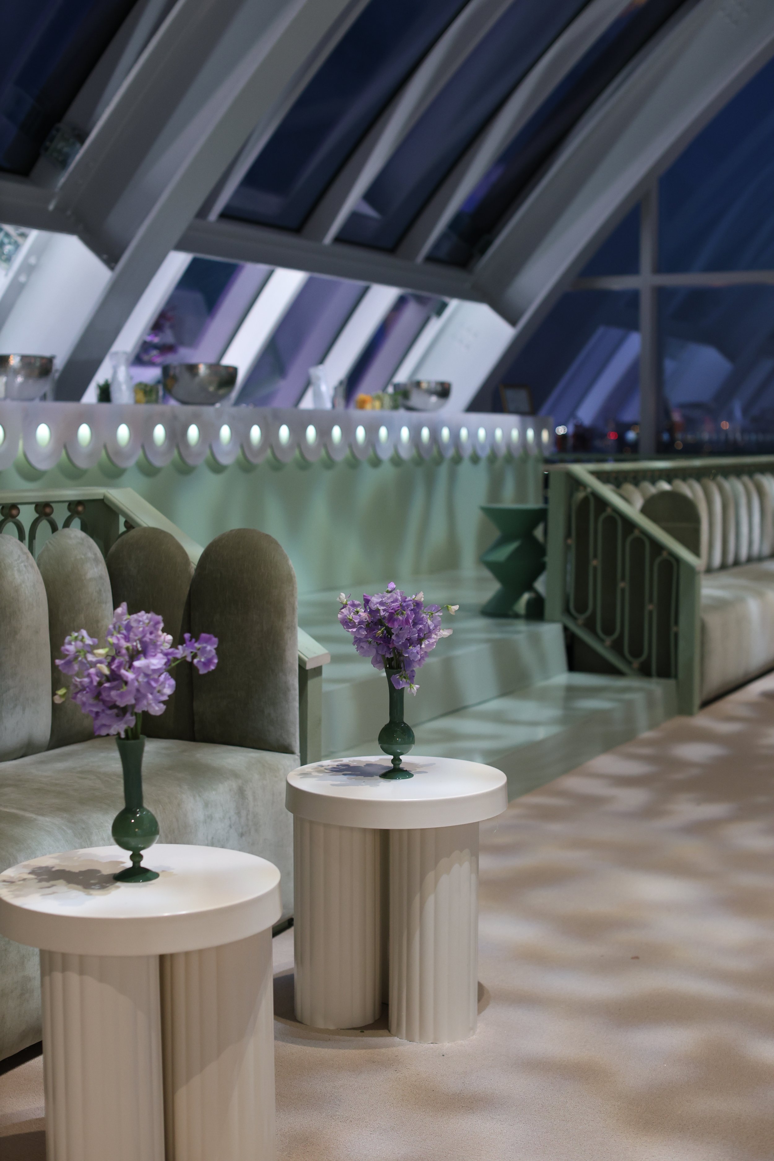 Interior of a modern, stylish room with a staircase, purple flowers on two small white tables, velvet blue chairs, and a hill of windows with a night sky outside.