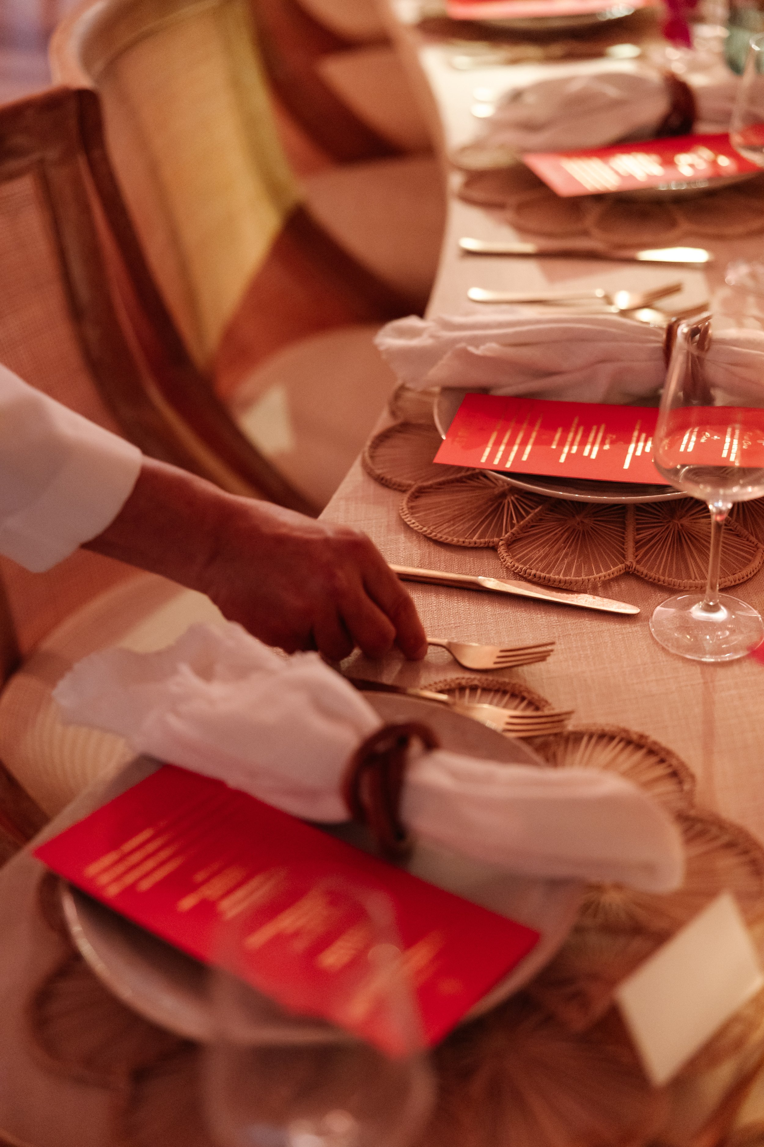 A long dining table set for a meal with place settings, napkins, and menus. A person is seen placing a utensil on the table.