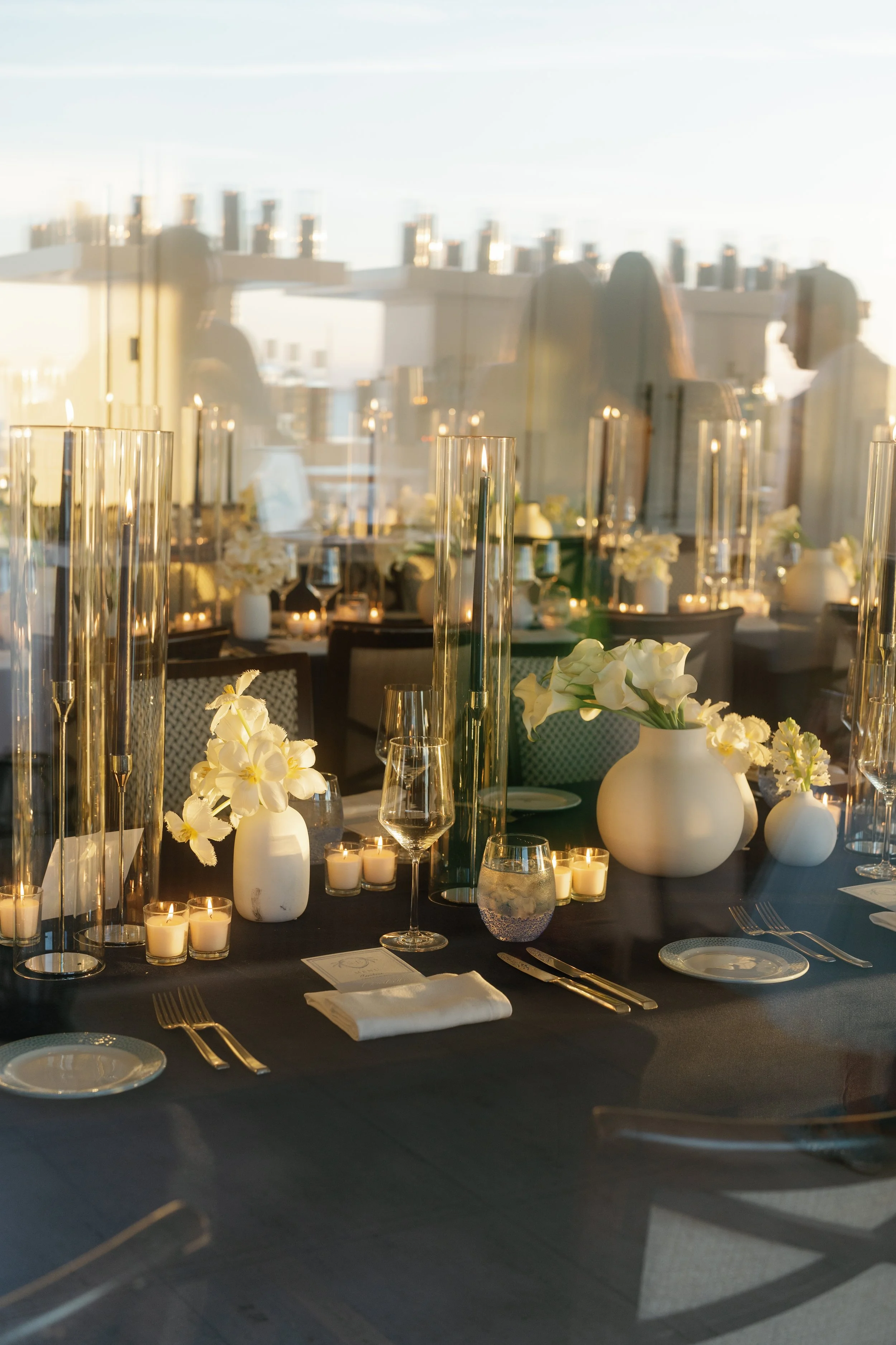 Elegant dining table set with white floral arrangements, candles, and glassware, seen through a window reflection, at a formal event or dinner.