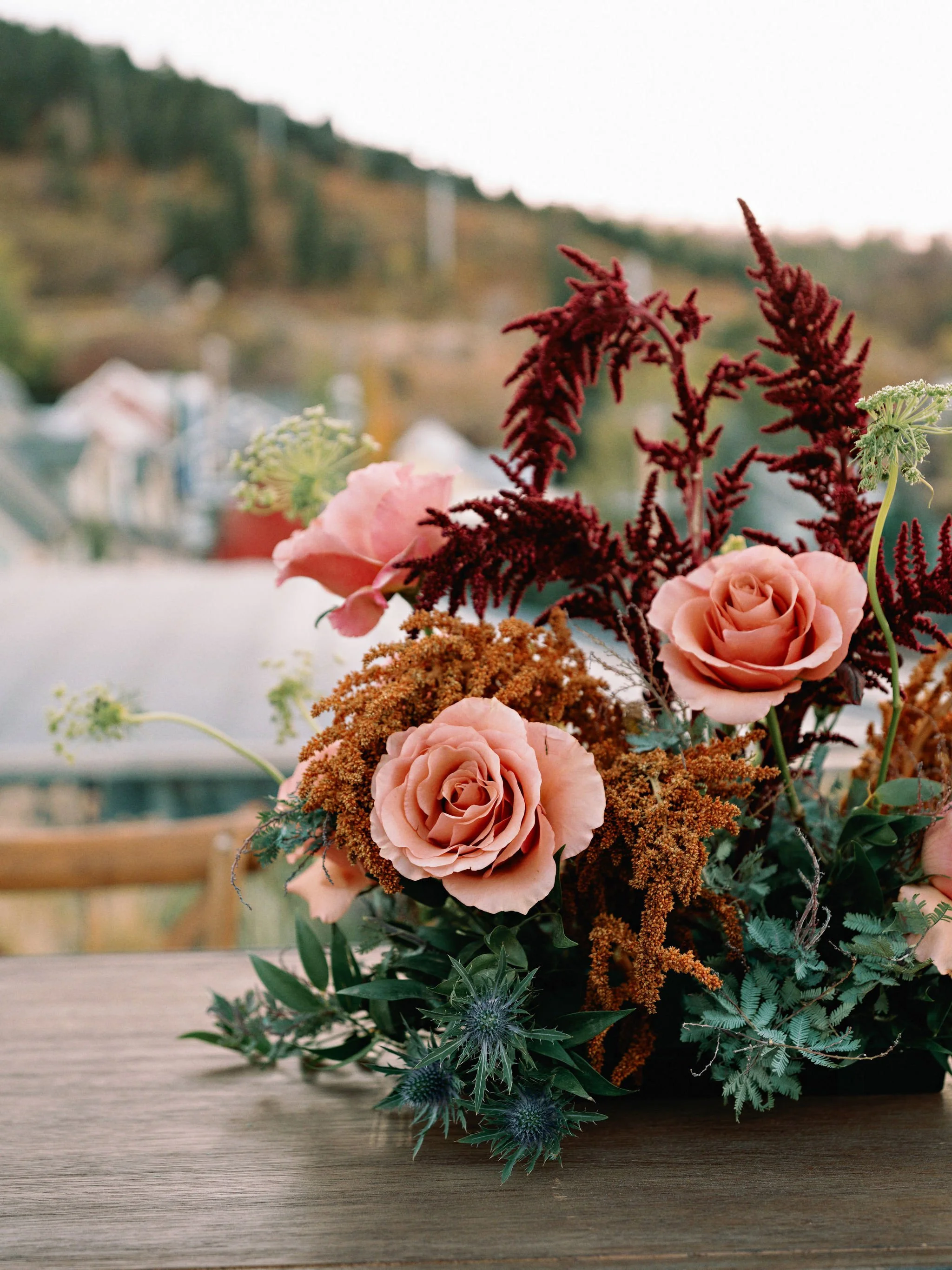 A floral arrangement with pink roses, red fern, brown and green foliage, and blue thistle flowers, set on a wooden surface with a blurred outdoor background.