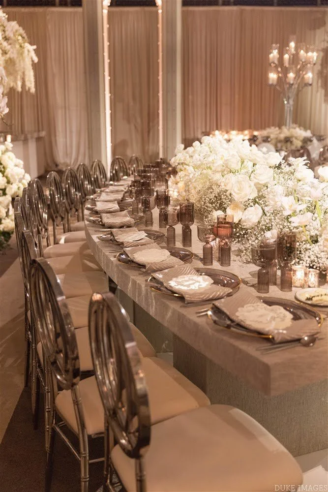 Elegant banquet table decorated with white floral arrangements, with place settings including plates, napkins, and silverware, and surrounded by metallic chairs with light-colored cushions, in a warmly lit indoor setting.