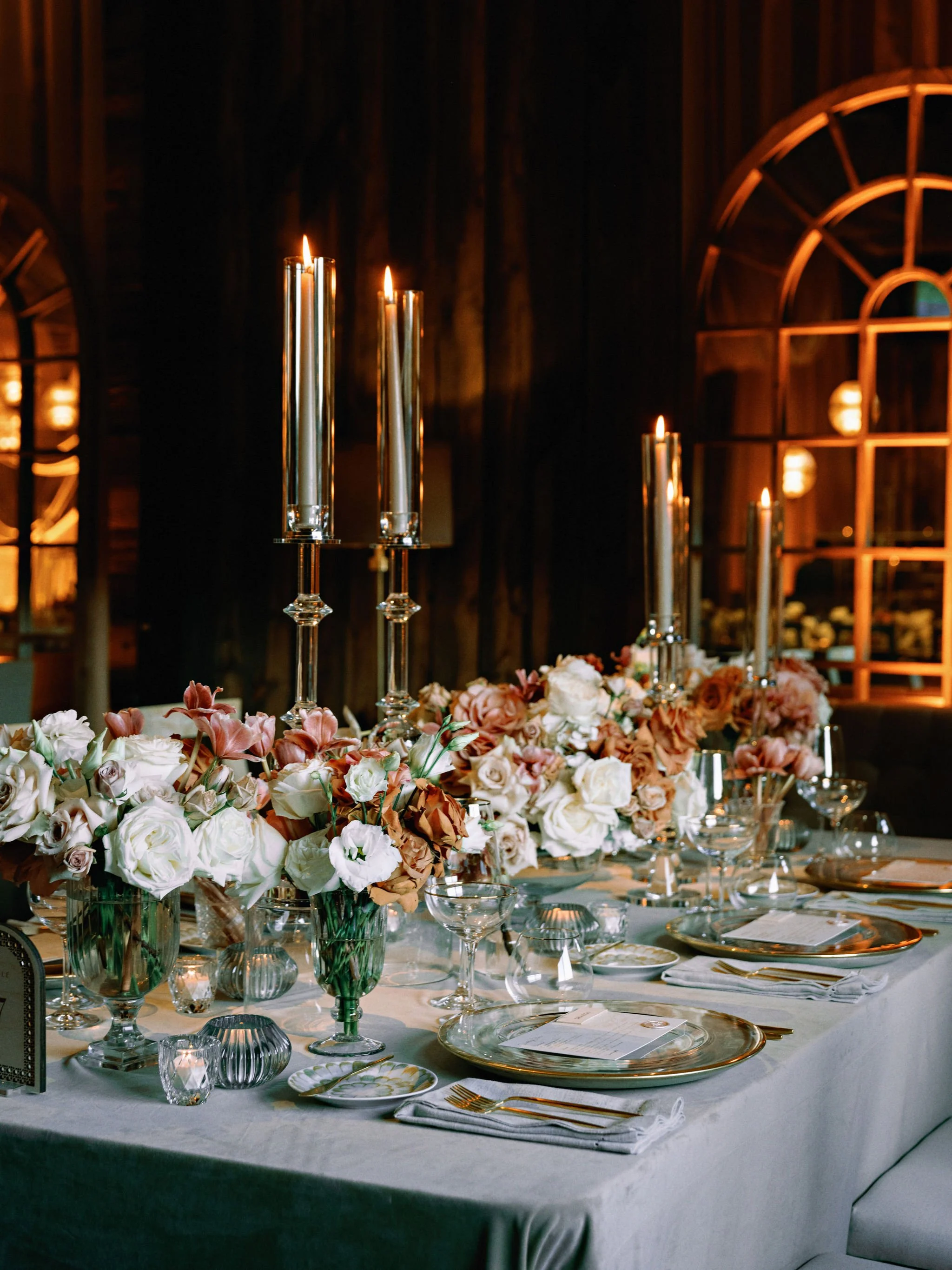 A beautifully decorated banquet table with floral centerpieces, tall candles, glassware, plates, and cutlery, set for a formal event in a dimly lit room with wooden walls and arched windows.