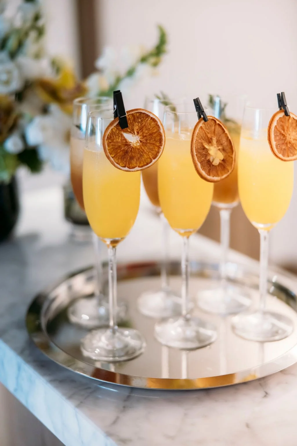 Five champagne flutes filled with a yellow beverage, garnished with dried orange slices attached to clothespins, arranged on a silver tray.