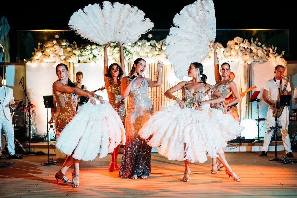 A group of performers, including women in white feathered skirts and gold dresses, singing and dancing on stage with musicians playing instruments in the background, at a lively event with floral and decorative backdrops.