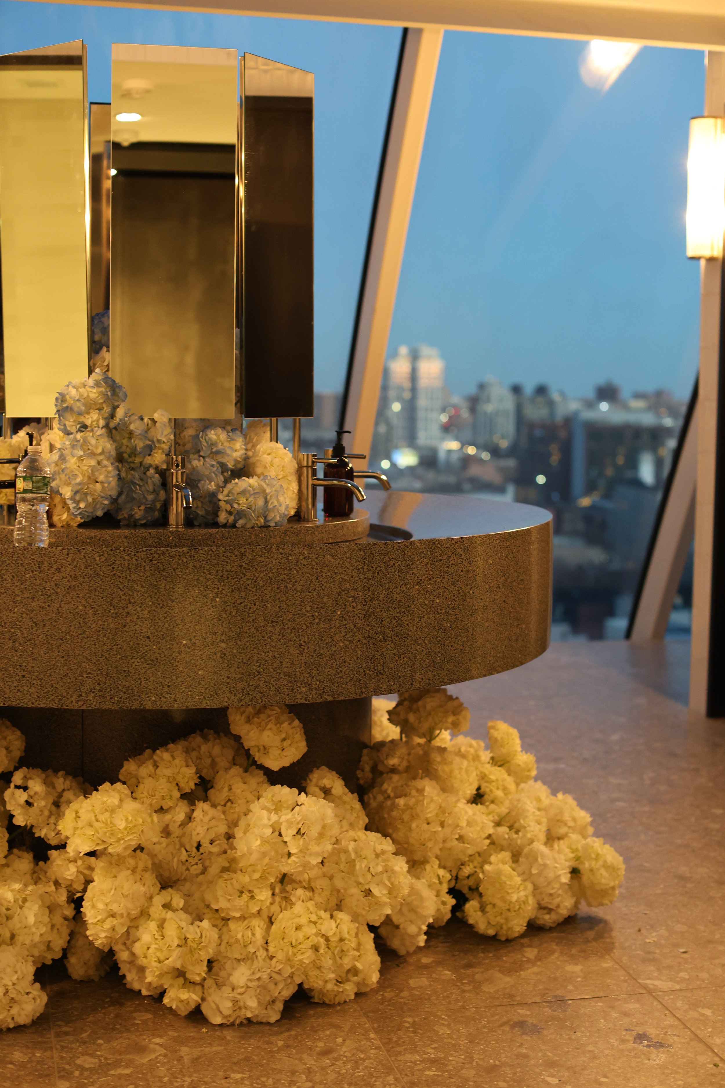 Wedding or event bathroom vanity decorated with white hydrangea flowers, with mirrors, soap dispenser, and hand sanitizer, located in a high-rise building with a city skyline view at dusk.