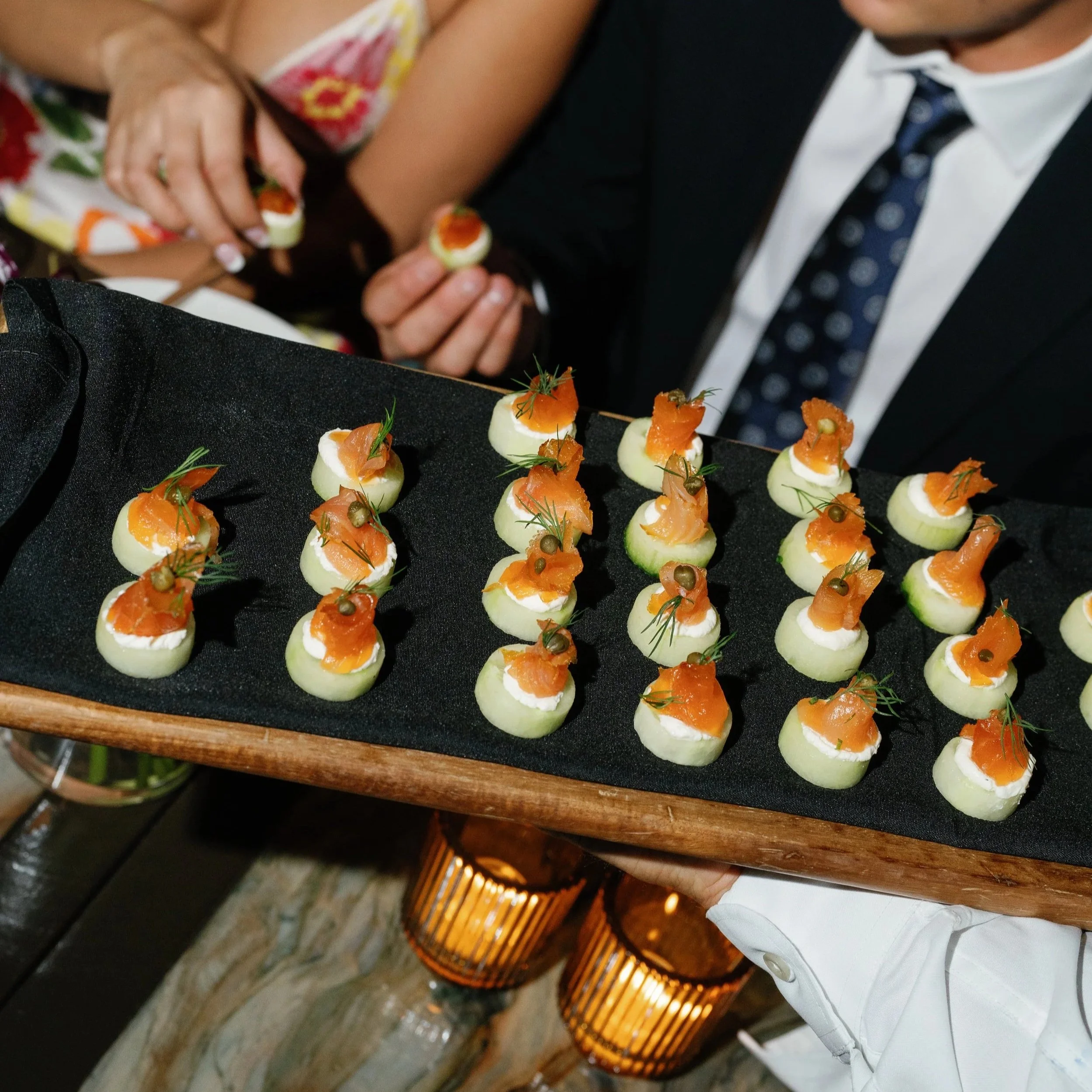 Tray of canapés topped with smoked salmon, dill, and capers on sliced cucumbers, served at a social event.