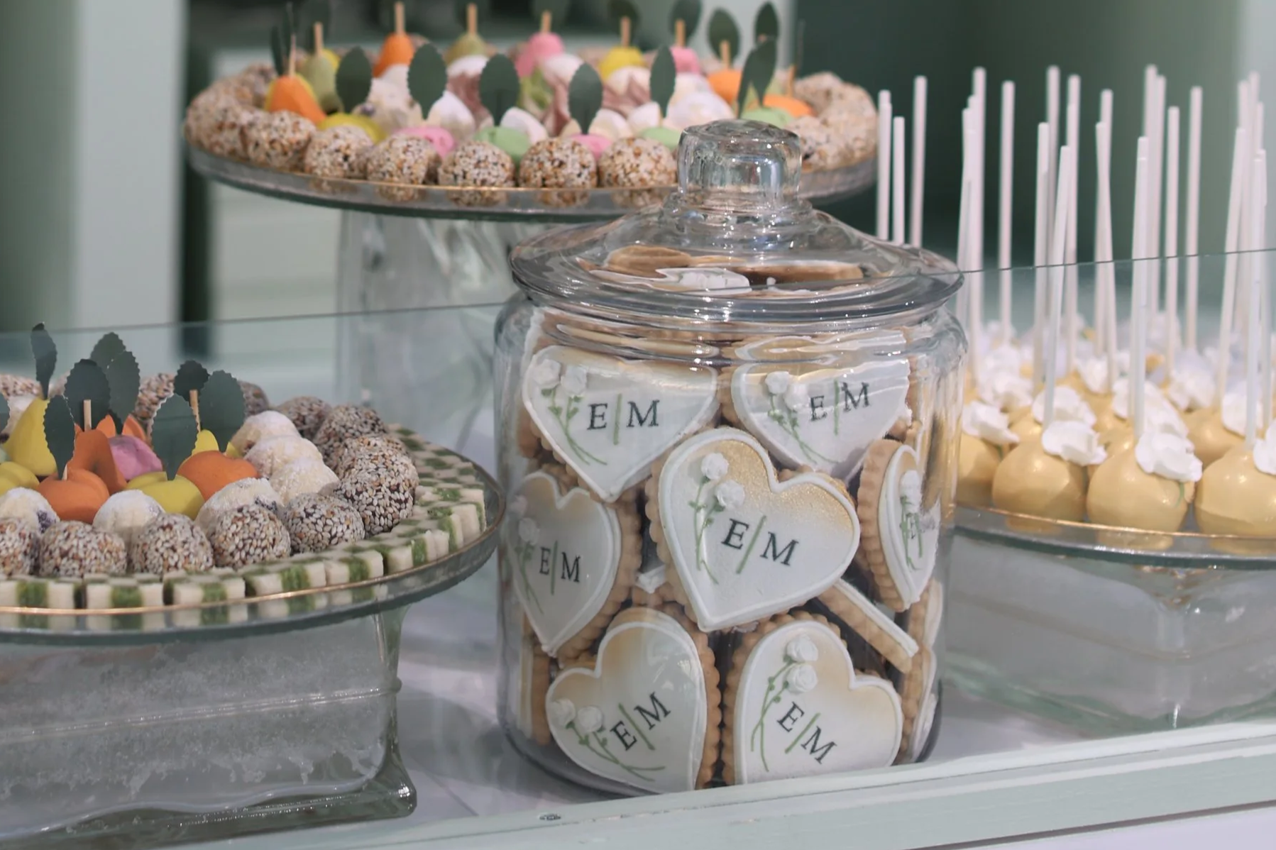 Assorted colorful candies and cookies displayed on a glass table, including decorated cookies with initials, candies on sticks, and a jar of heart-shaped cookies with initials.