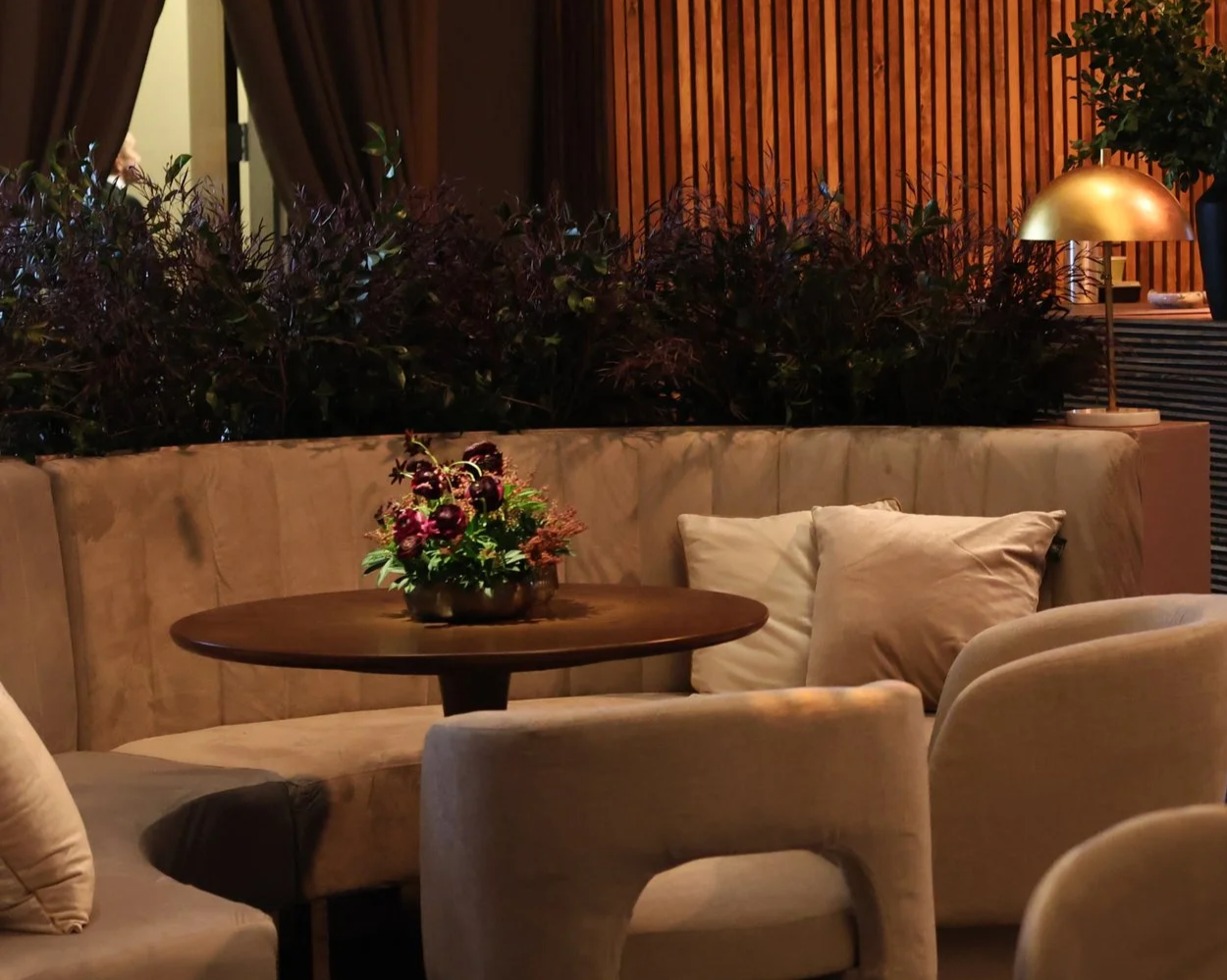 Indoor lounge area with beige sofa, round wooden table with flower arrangement, pillows, plants, and warm lighting.