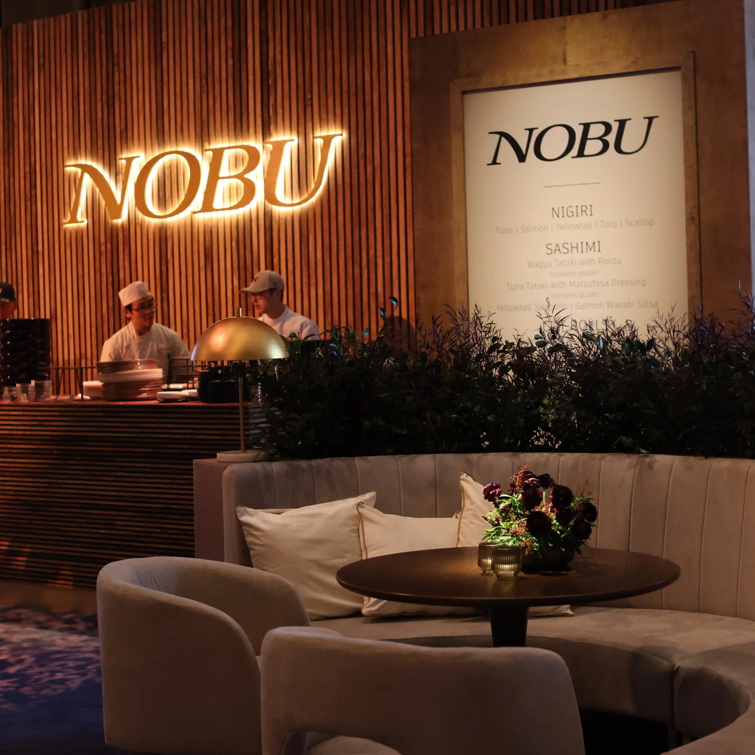 Interior of a restaurant with a wooden wall, illuminated 'NOBU' sign, a Japanese chef and worker behind the counter, a round table with a flower arrangement, and cushioned seating.