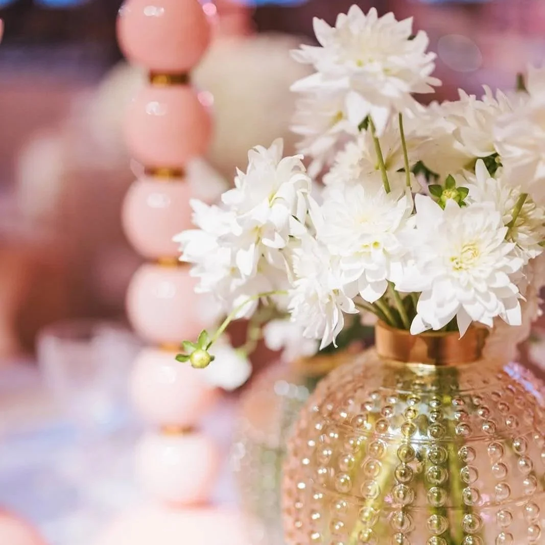 White flowers in a decorative pink and gold vase with soft-focus pink and gold objects in the background.