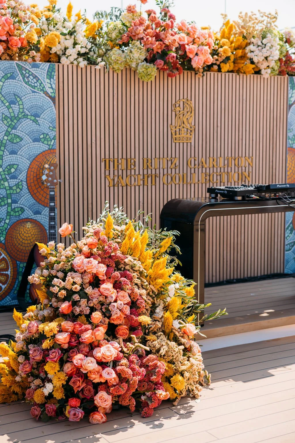 Decorative stage setup with a large bouquet of pink, yellow, and white flowers, a wooden backdrop with gold lettering that reads 'The Ritz Carlton Yacht Collection,' a DJ booth, and colorful mosaic art on the sides.