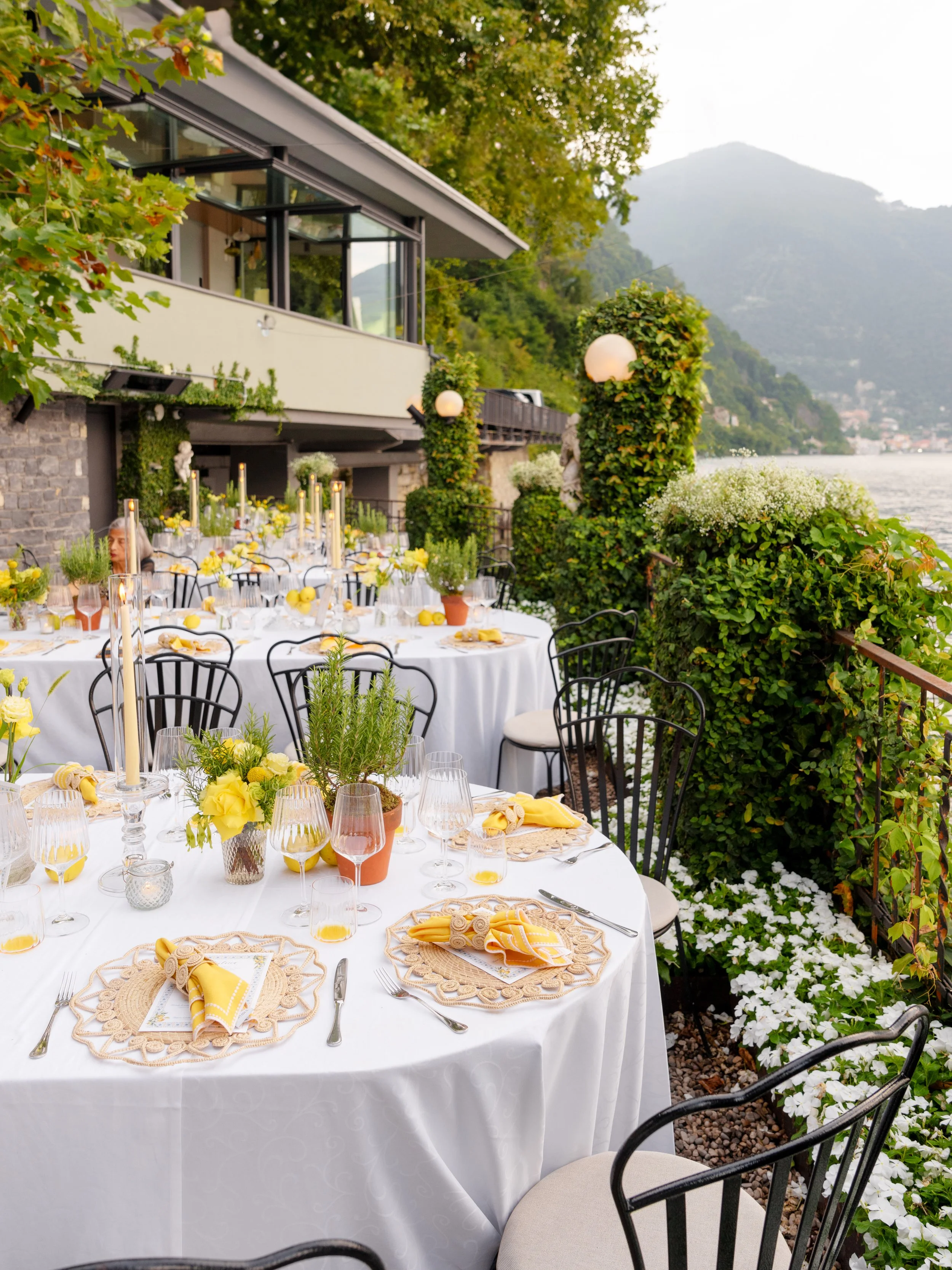 Elegant outdoor dining setup on a balcony overlooking a lake, with white round tables covered with white tablecloths, decorated with yellow flowers, candles, and placemats with yellow napkins, surrounded by black chairs with cream seats, green plants