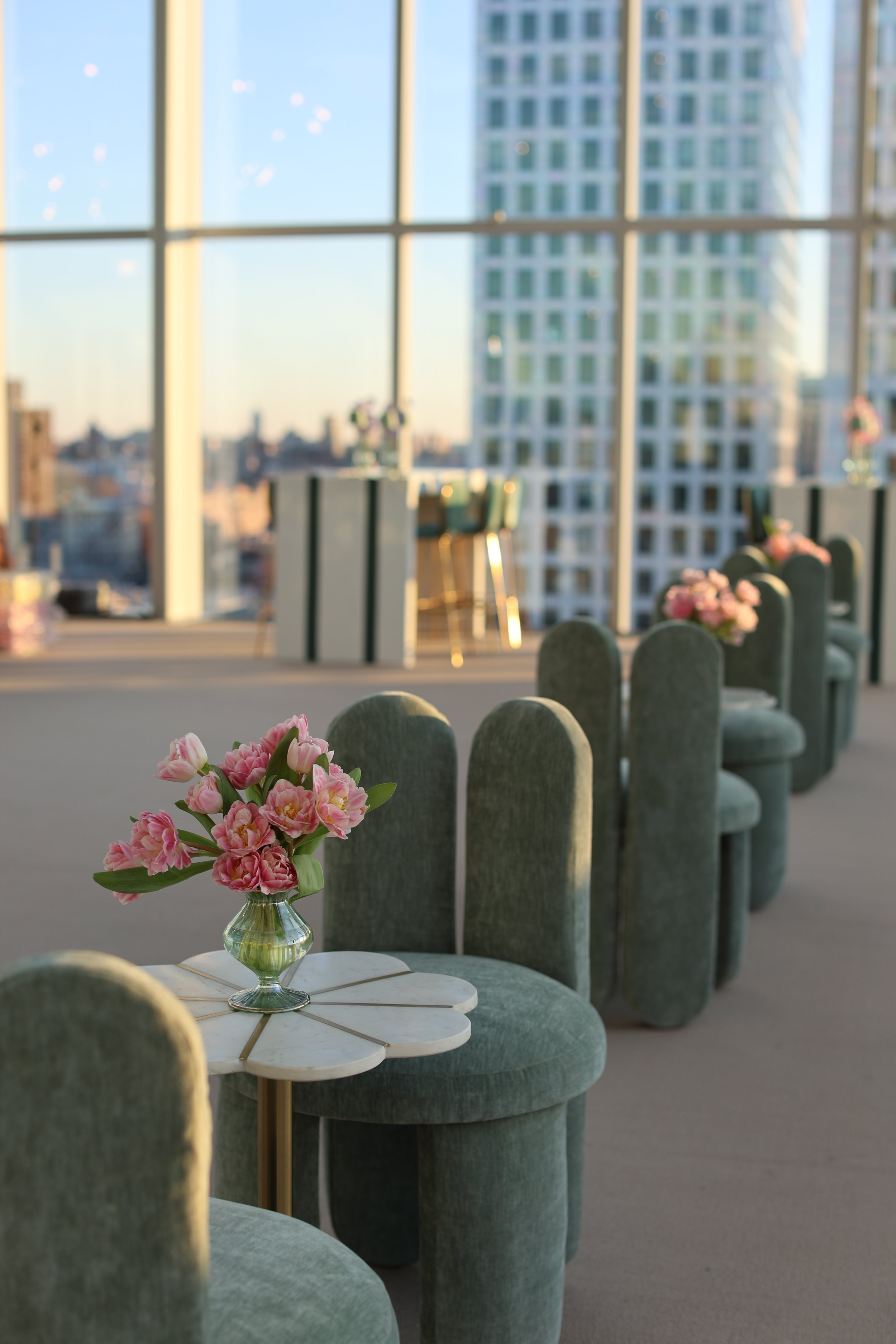 Interior of a modern high-rise apartment with green velvet chairs arranged around small tables with pink flowers, large windows with city skyline views, during sunset.