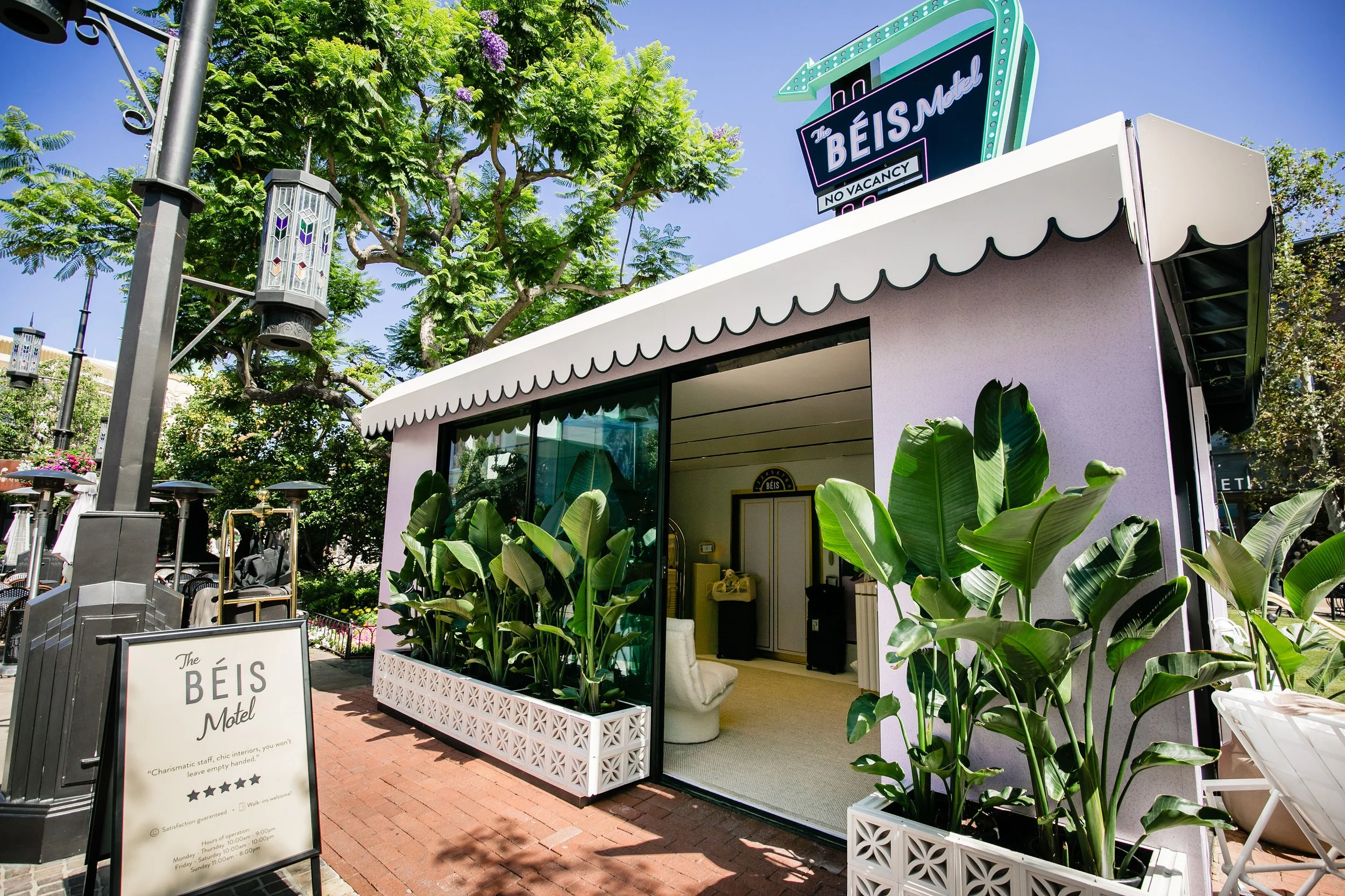 Exterior view of The Beis Motel with a pink facade, large green plants in white decorative planters, and a signboard outside. The sign says 'The BEIS Motel' and includes a rating of five stars. The interior glimpse shows a cozy seating area with a wh