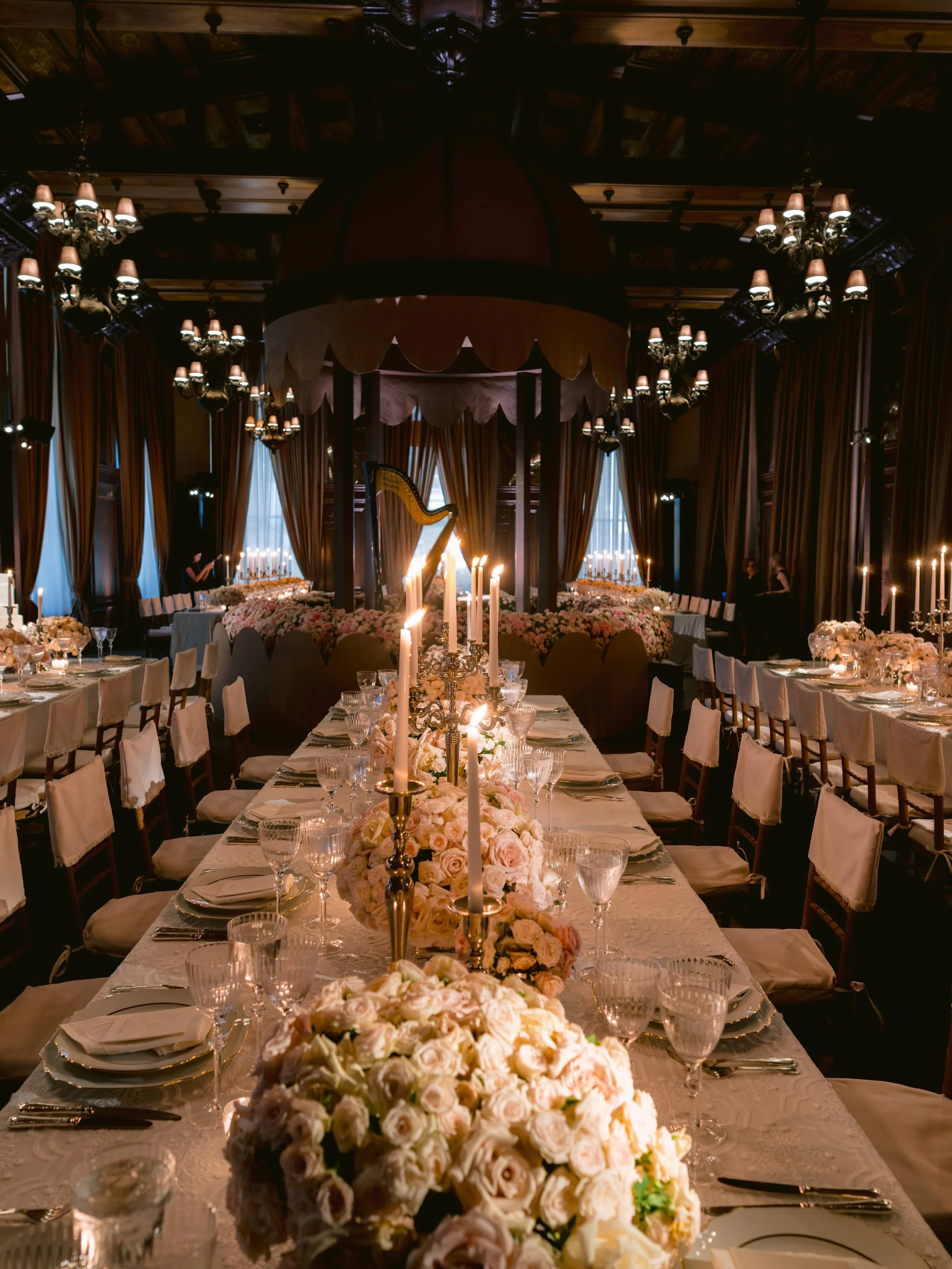 Elegant banquet table set with floral centerpieces, white tablecloths, fine glassware, and candles, in a luxurious decorated room with chandeliers and draped curtains.