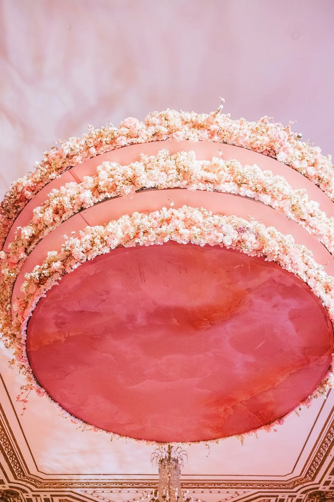 Decorative ceiling with pink circular panels bordered by white and pink flowers, ornate gold trim, and a chandelier hanging below.